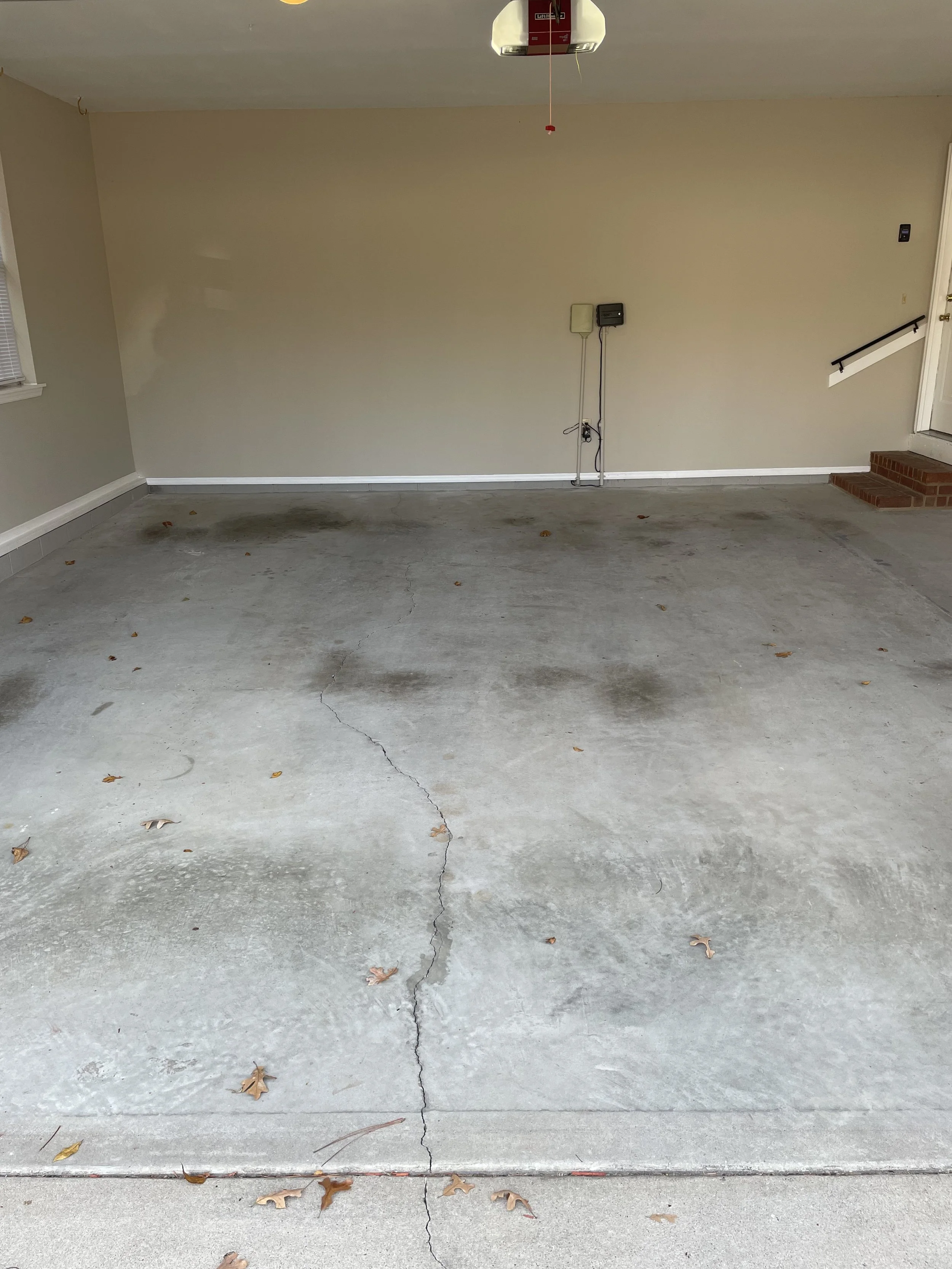 Empty garage with a cracked concrete floor, beige walls, and some leaves scattered on the ground.
