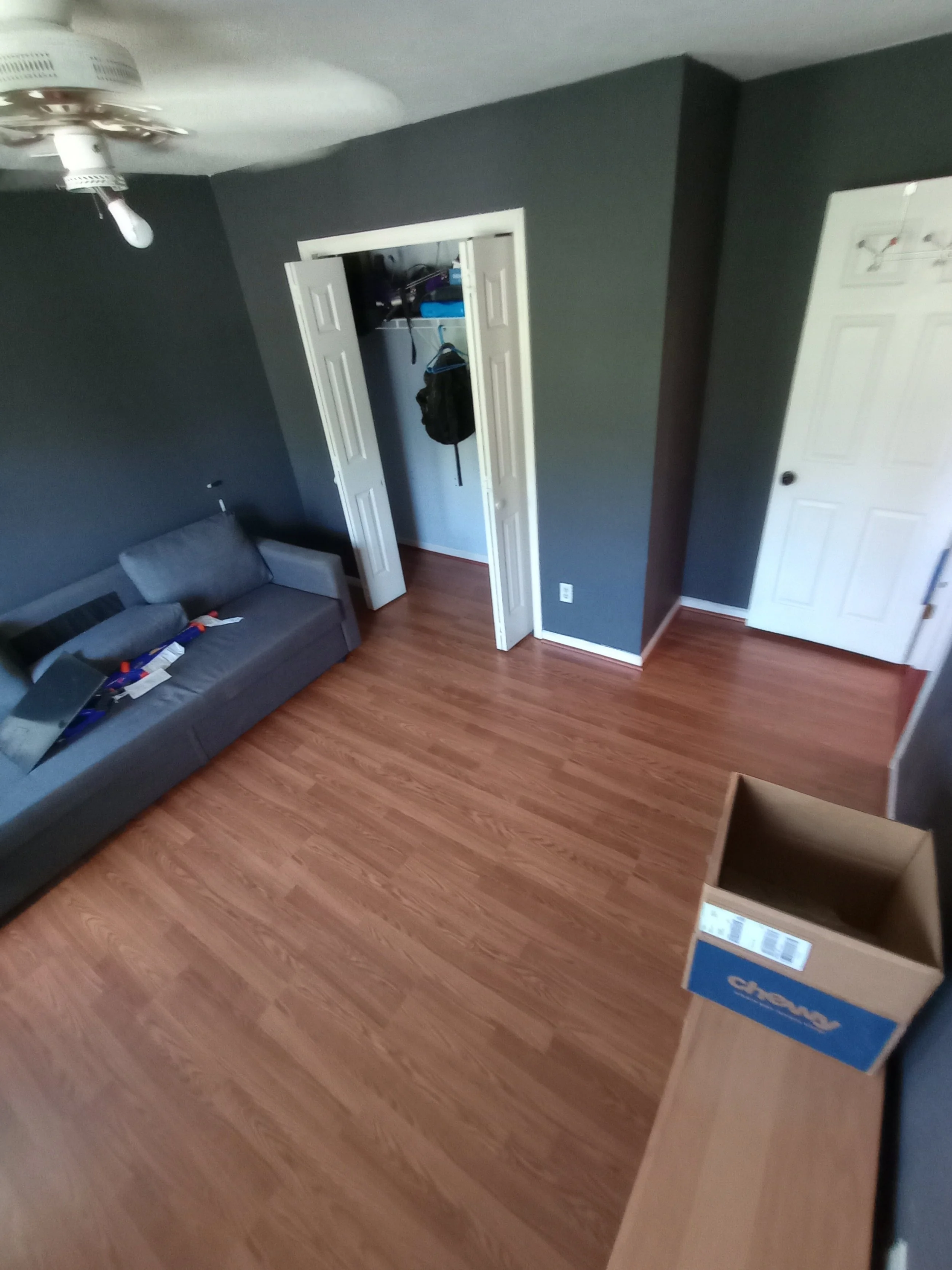 Empty room with a gray sofa, a closet with open double doors, a cardboard box on a wooden table, and a ceiling fan with light.