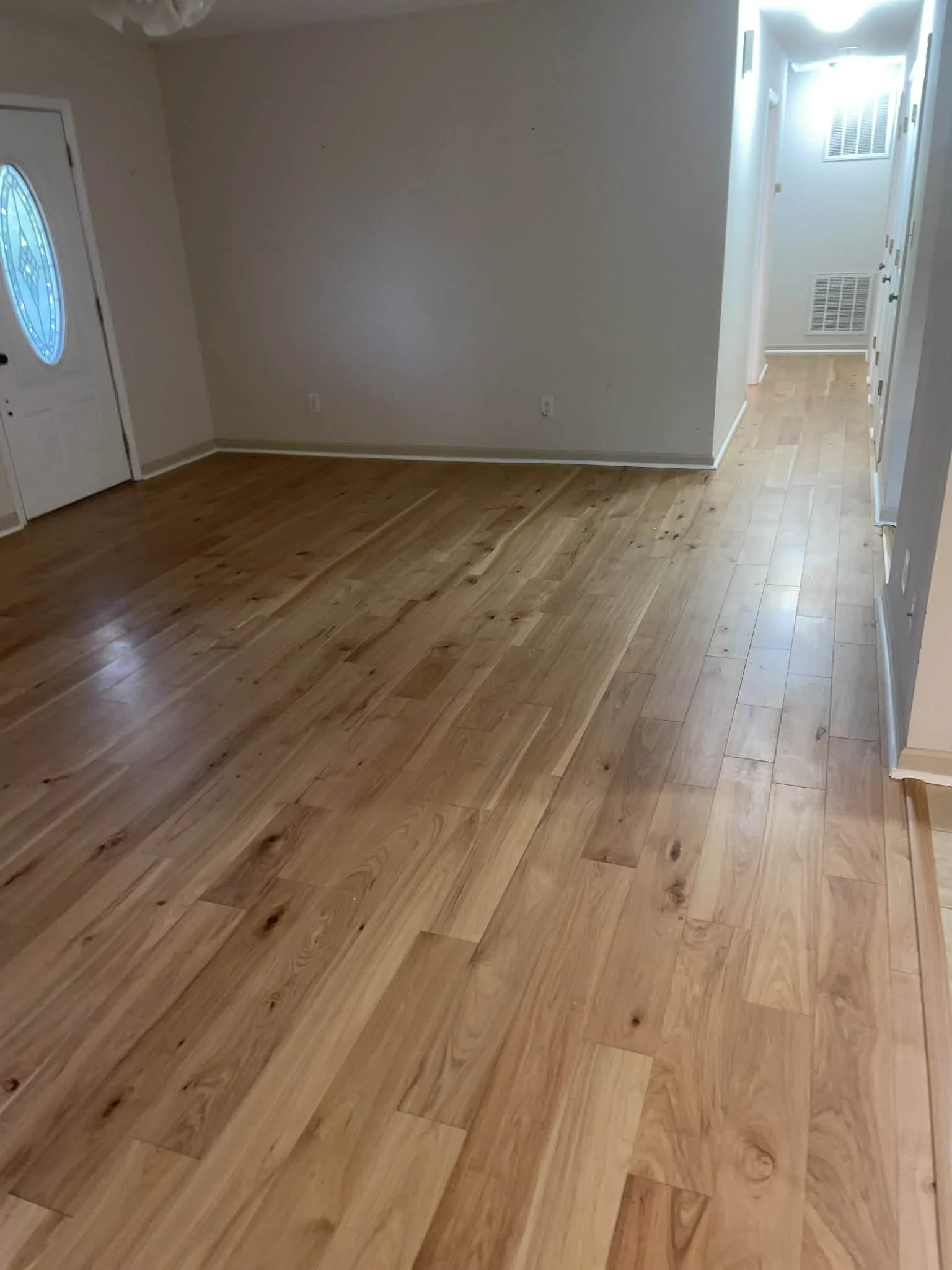 Empty living room with hardwood flooring, closed front door with a decorative glass window, and a hallway leading to other rooms.
