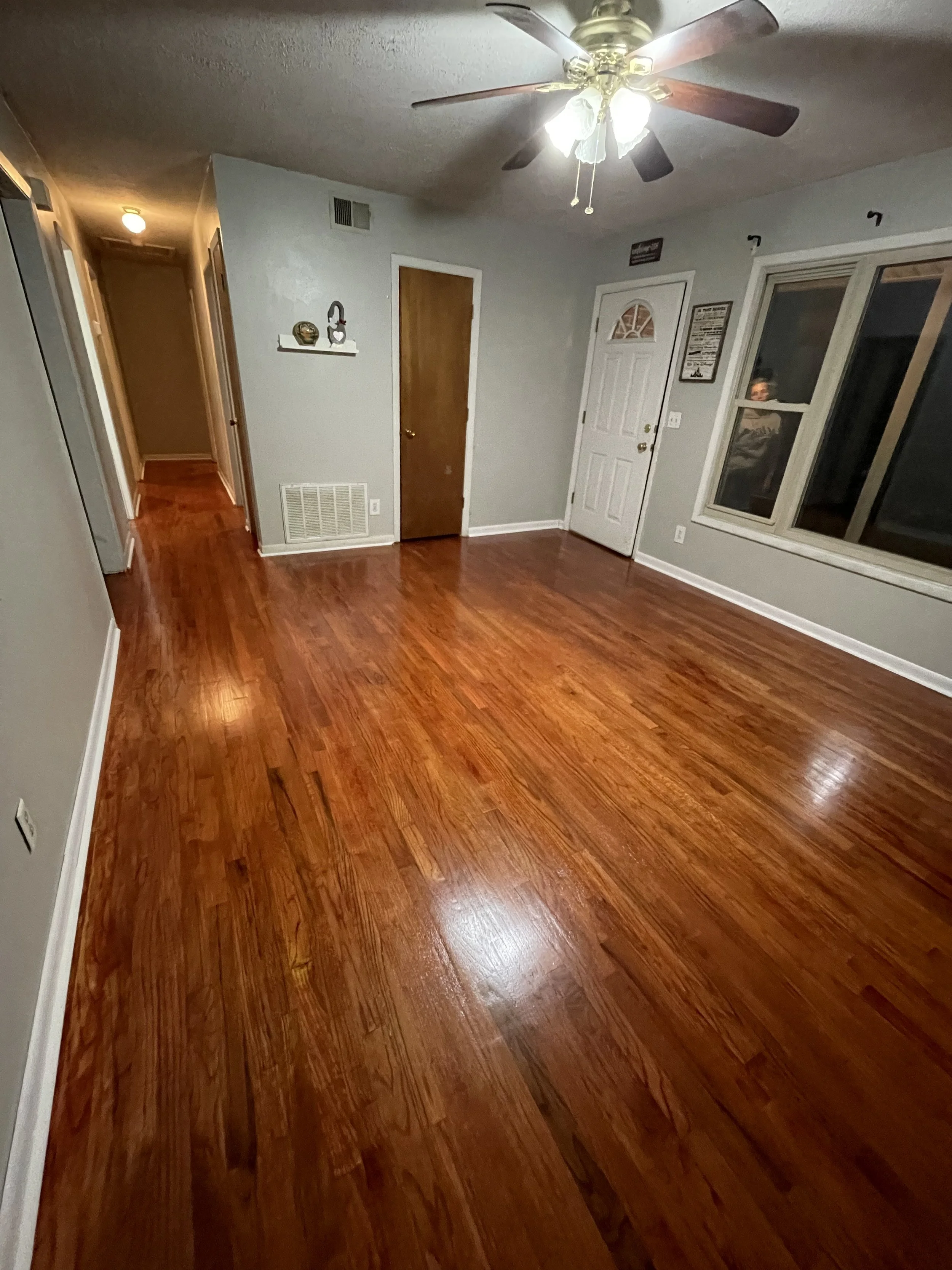 Empty living room with polished hardwood floors, a ceiling fan with lights, a large window, and two doors.