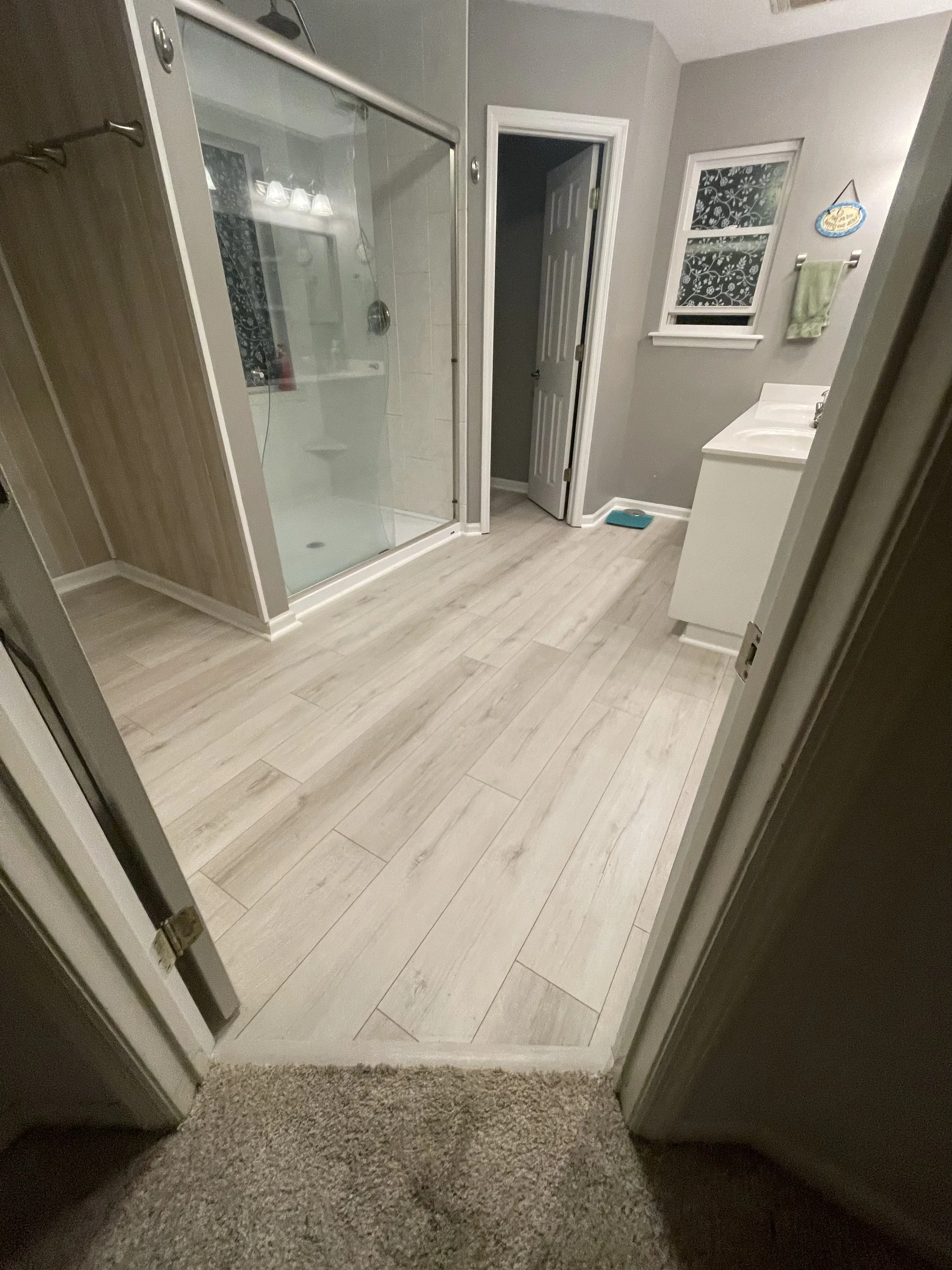 View of a bathroom with a walk-in shower with a glass door, a small window with floral curtain, a vanity with a sink, a towel hanging on a rack, a scale on the floor, and a door leading to another room.
