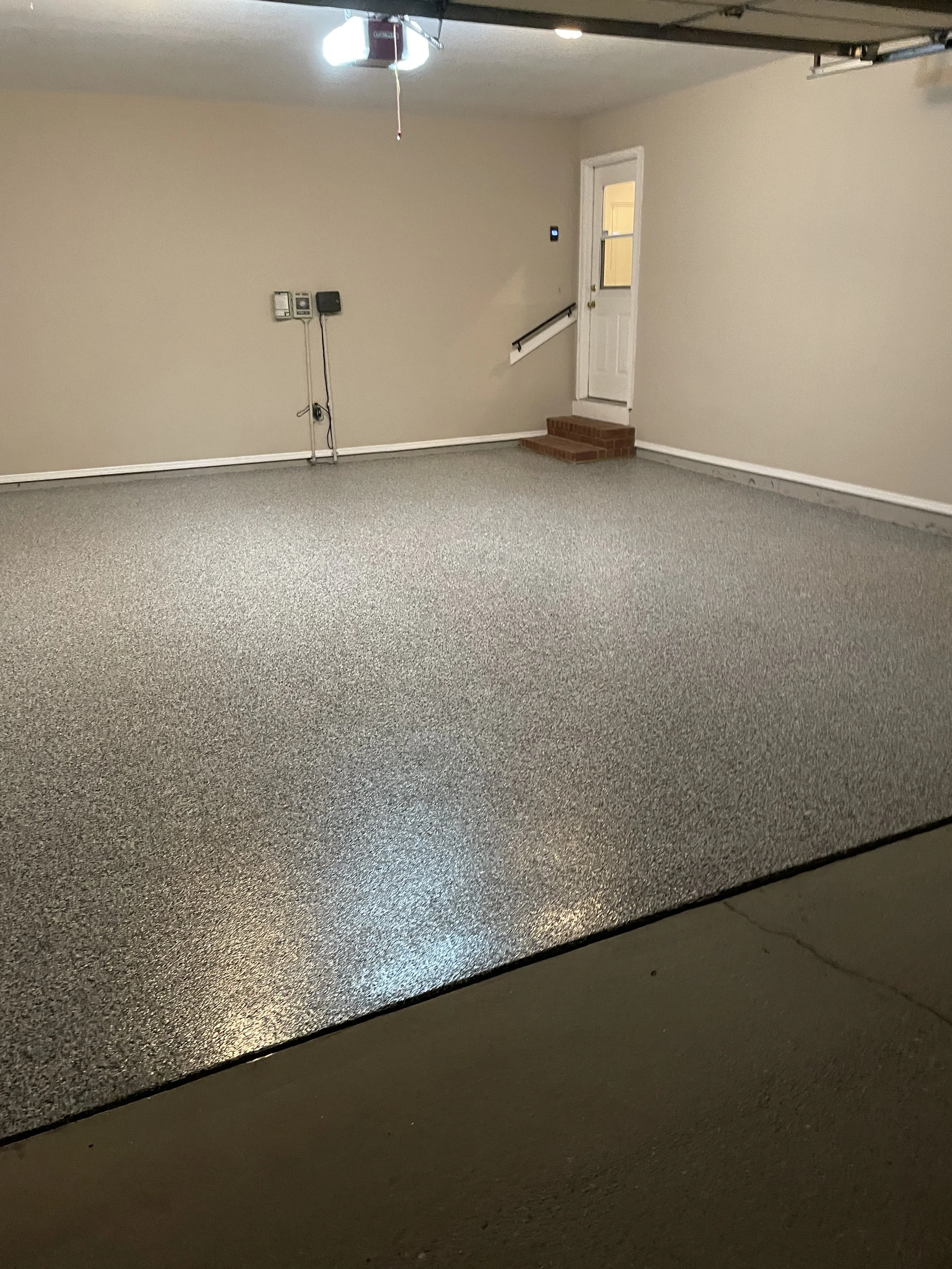 Empty garage with beige walls, gray textured floor, white trim, a door with a small window, a staircase with brick steps, and electric outlets on the wall.