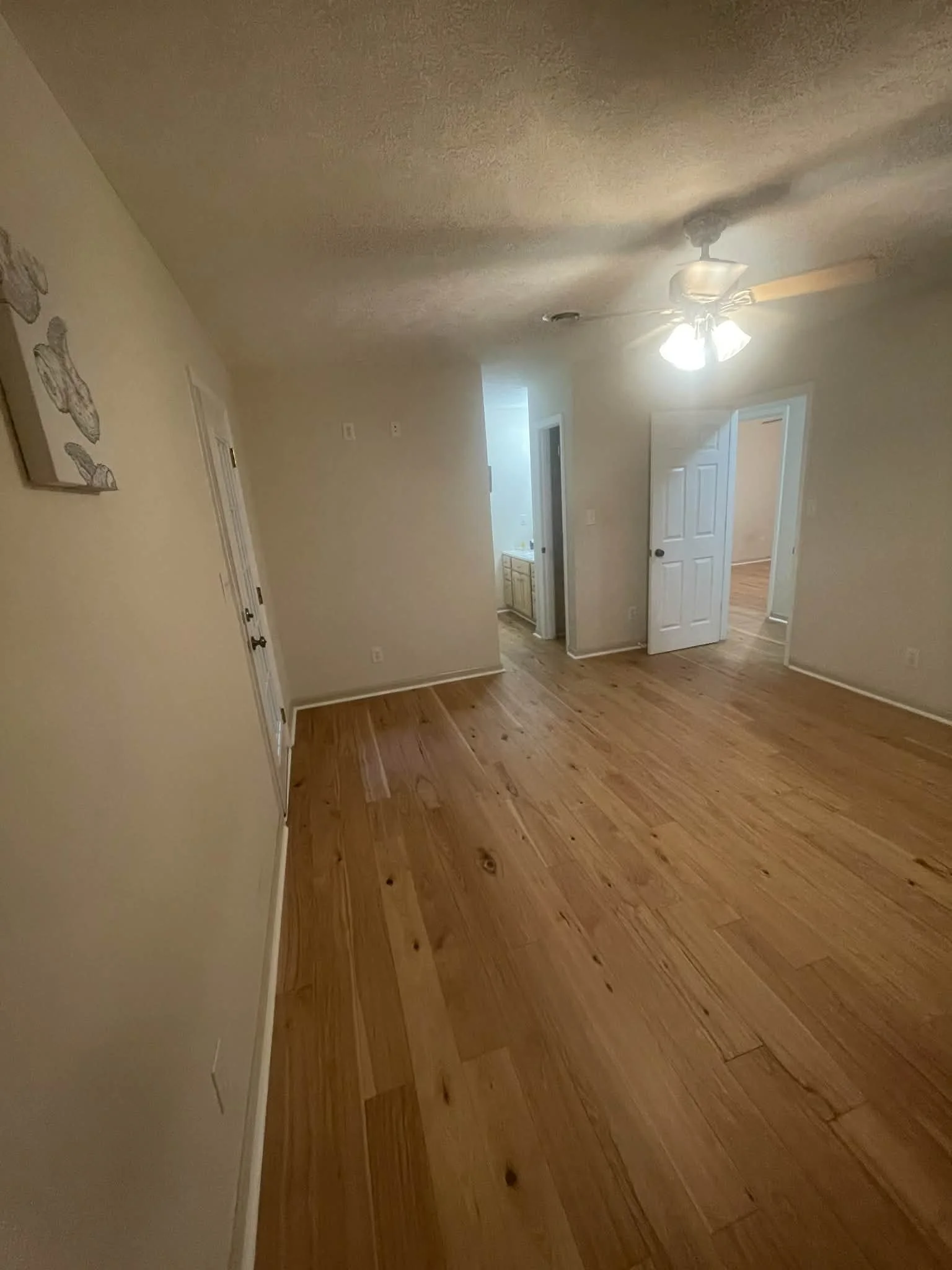 Empty living room with wooden floor, ceiling fan with lights, open doorway to another room, and beige walls.