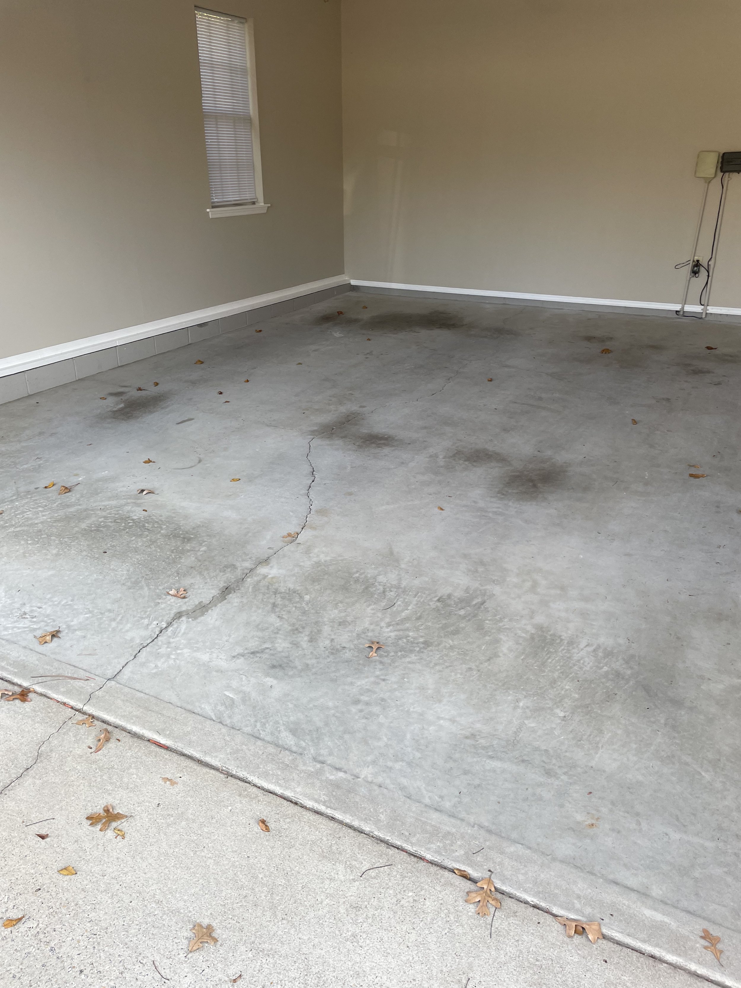 Empty garage with stained concrete floor, some leaves scattered, beige walls, and a small window with blinds.