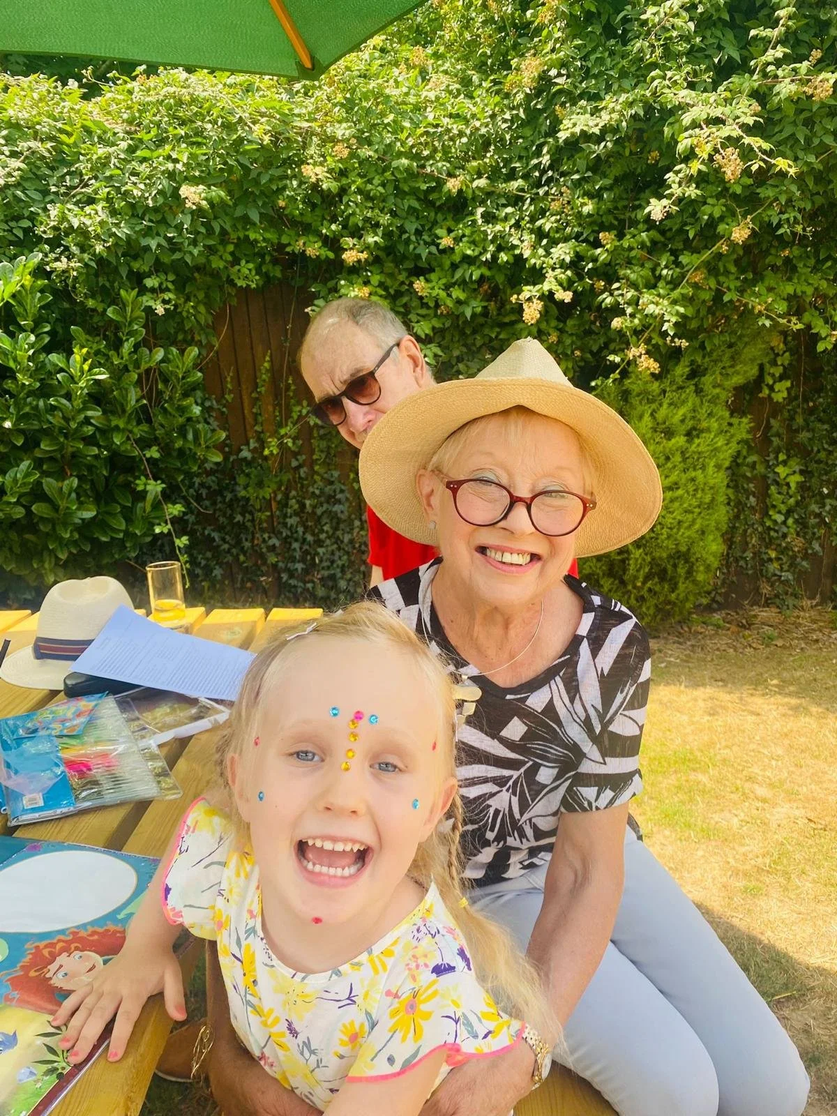 A vibrant and happy Jenny (living with HER2 lung cancer) with a grand daughter on a sunny day.  They're at a picnic table and Jenny is wearing a straw hat and sunglasses. The granddaughter is smiling and wearing stickers on her face.