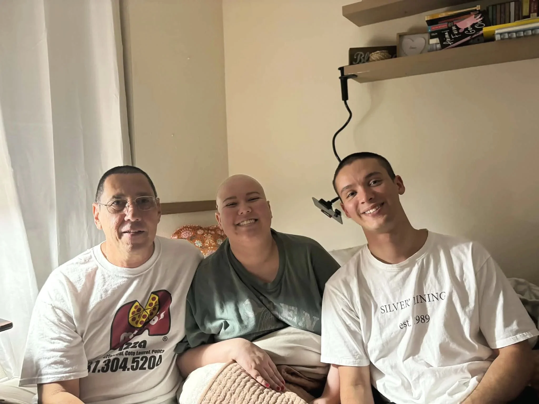 A young Camila, age 25 sits bald on a hospital bed next to her dad and younger brother as she is undergoing chemo and treatment for her KRAS G12V lung cancer.