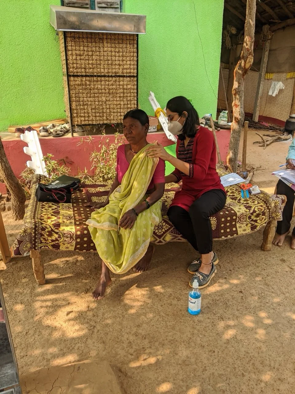 Rasika, in her twenties working as a physiotherapist in India. She wears a fight pink top and is treating a woman in a sari against a bright green wall in what appears to be a rural village.