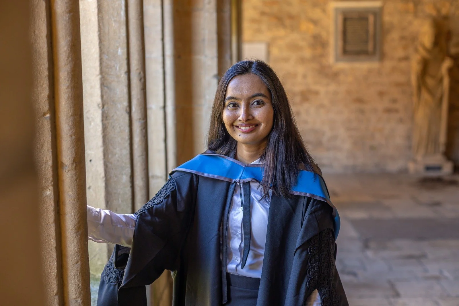 A smiling, young Raskia wearing her Oxford University graduation gown and clothing with the uni background is smiling at graduation - post her lung cancer diagnosis