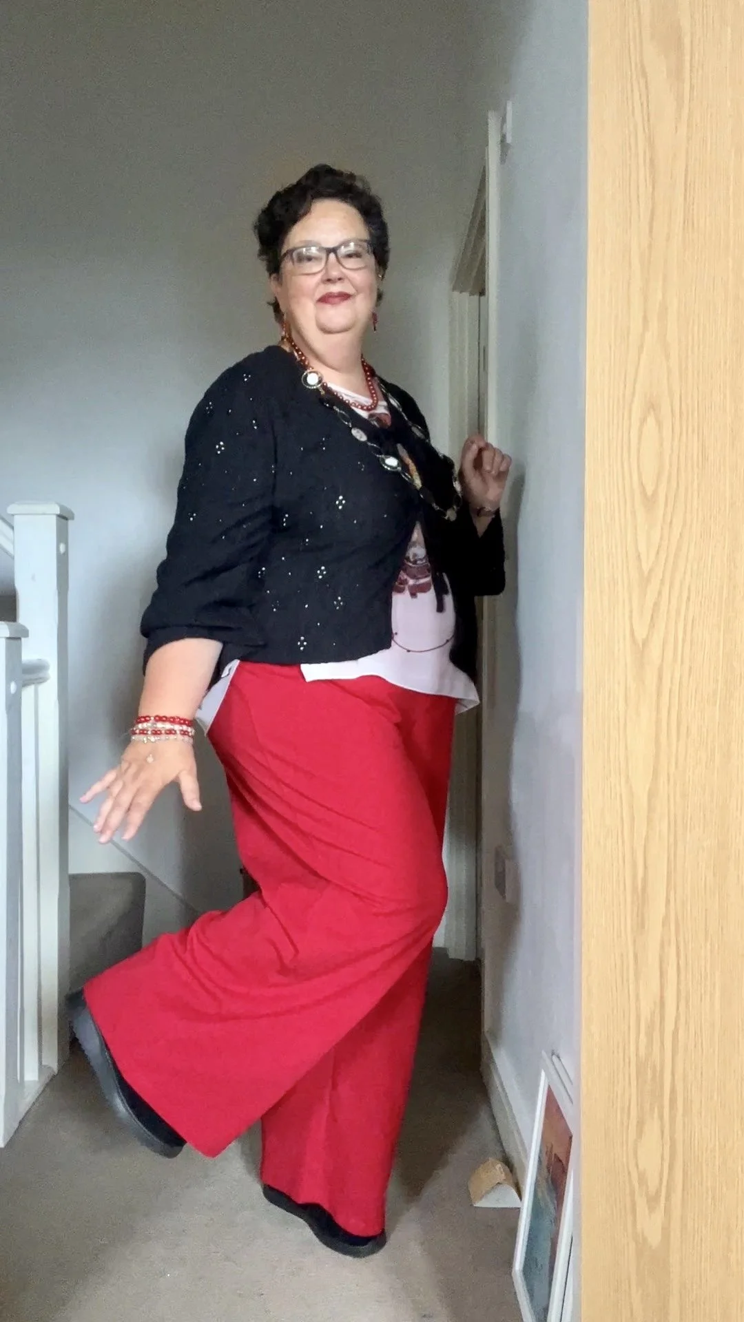 Allison by her staircase wearing bright red trousers and red lipstick, looking happy.
