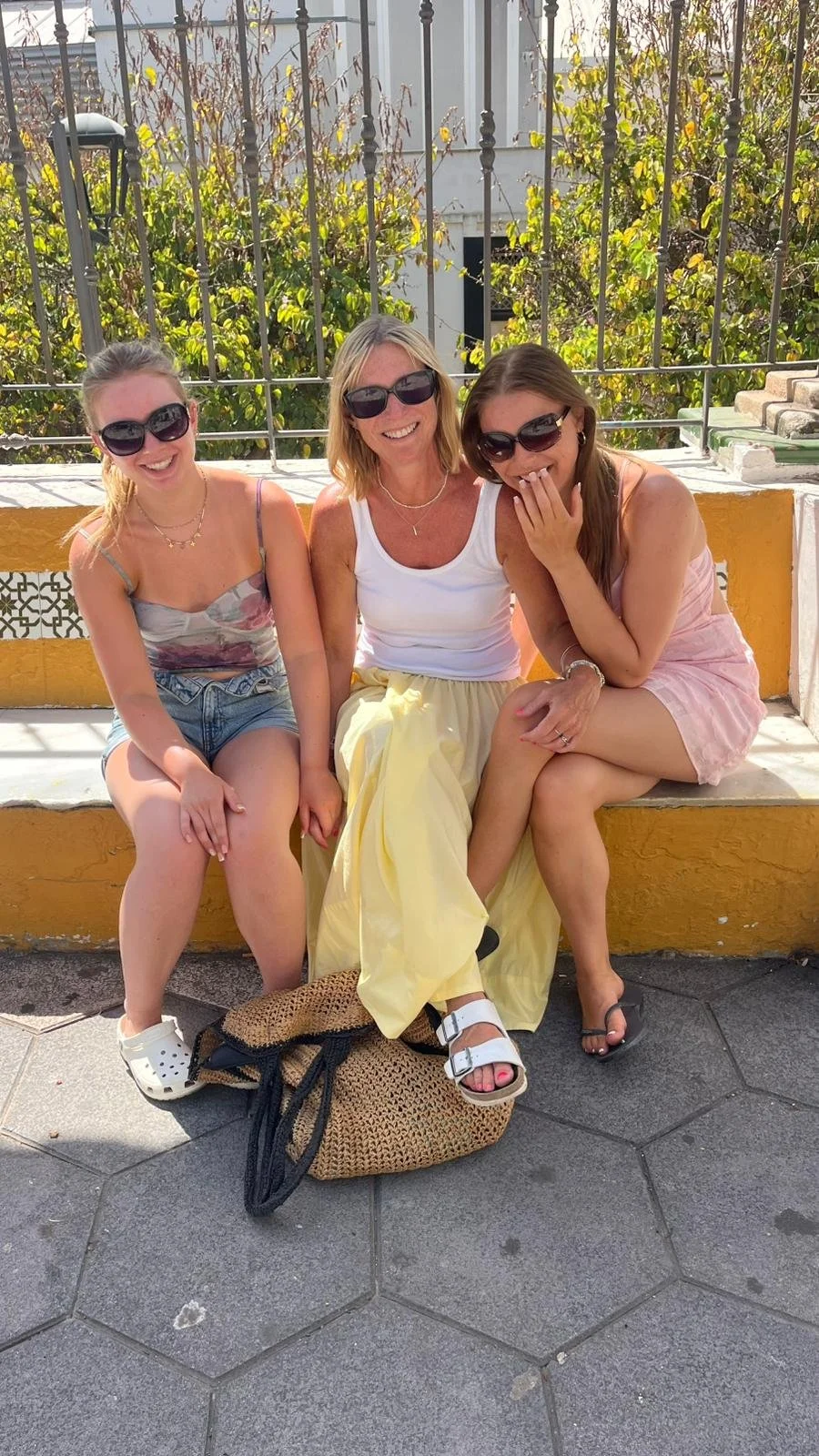 Natasha sits in between her two young teenage daughters.  All smiling and wearing sun glasses, shorts, skirts in a warm and sunny location.
