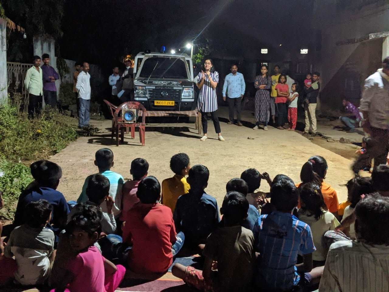 Rasika speaking in front of a large group of children in rural India at a microphone. Doing her health education.