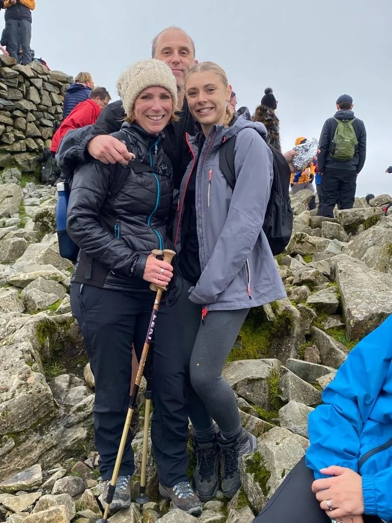Photo of Natasha, wearing a wooly hat, and holding a walking stick with her husband and a teenage daughter - climbing a very rocky hill.