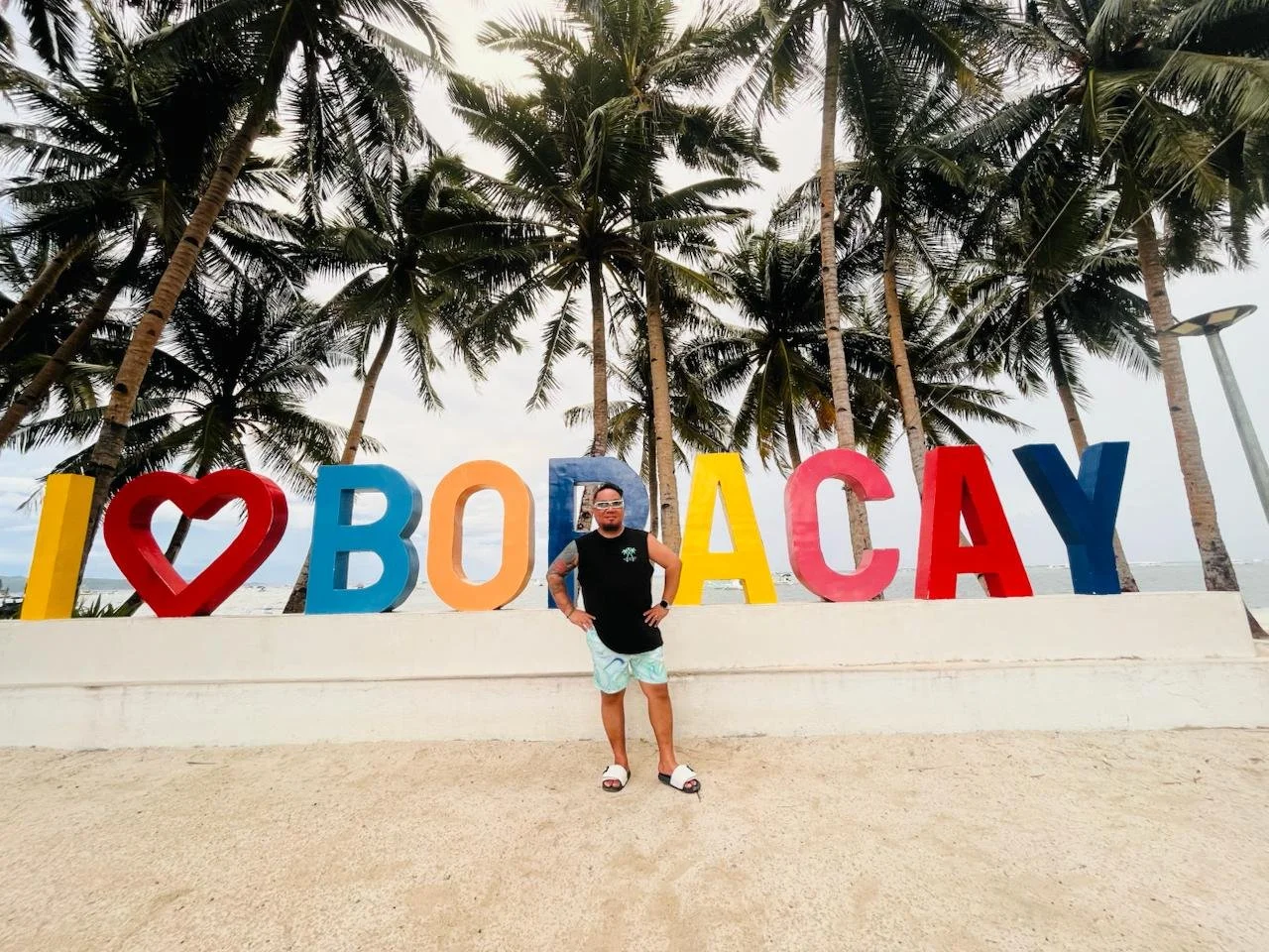 Tristan stands on a sandy beach, with big palm trees behind him and the ocean in front of a sign that says I love (heart sign) Boracay.  He stands with his hands on his hips and a big smile
