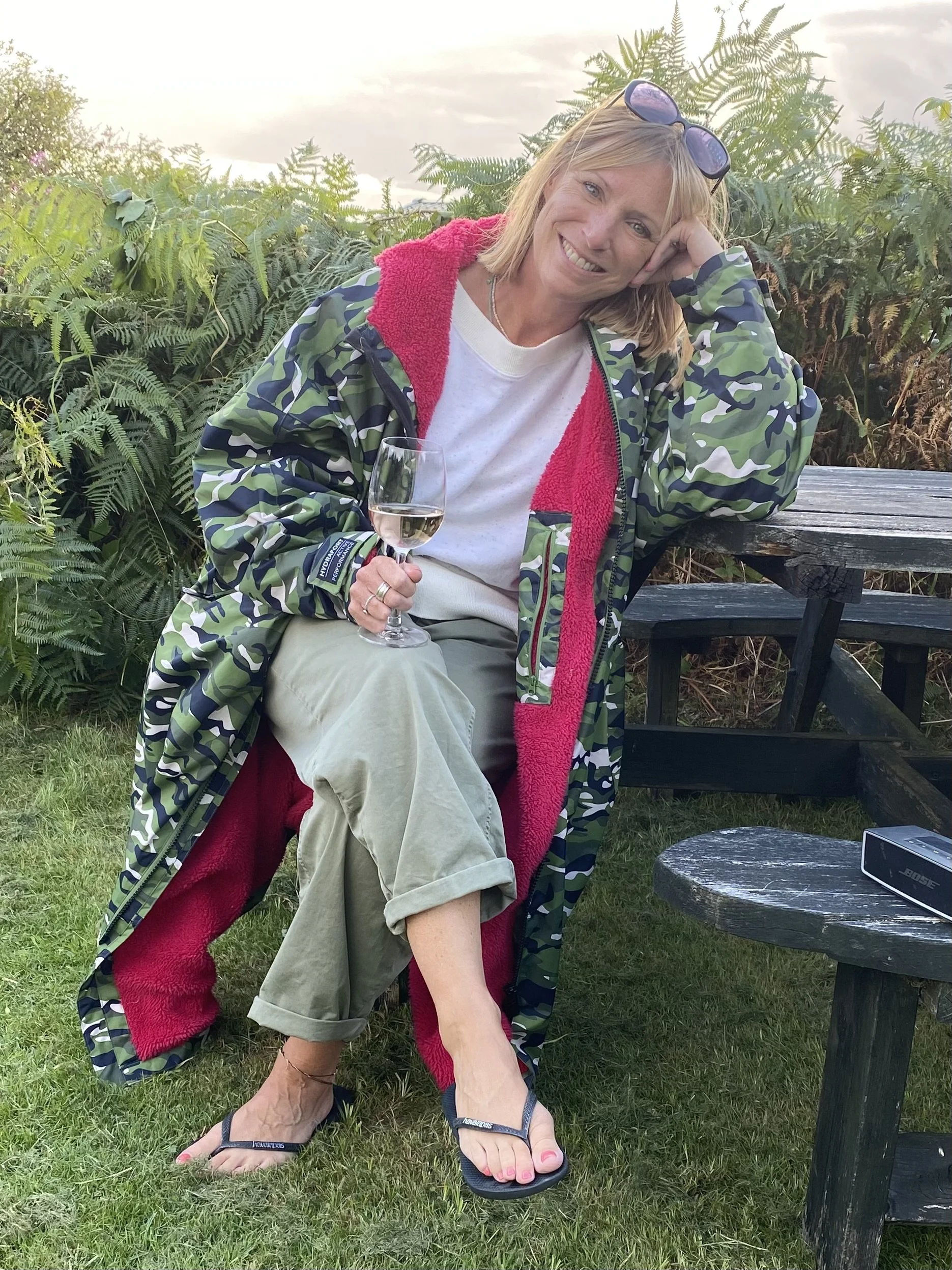 Natasha, sitting in a very green garden, wearing a camo long jacket with a big smile and a glass of wine in her hand.