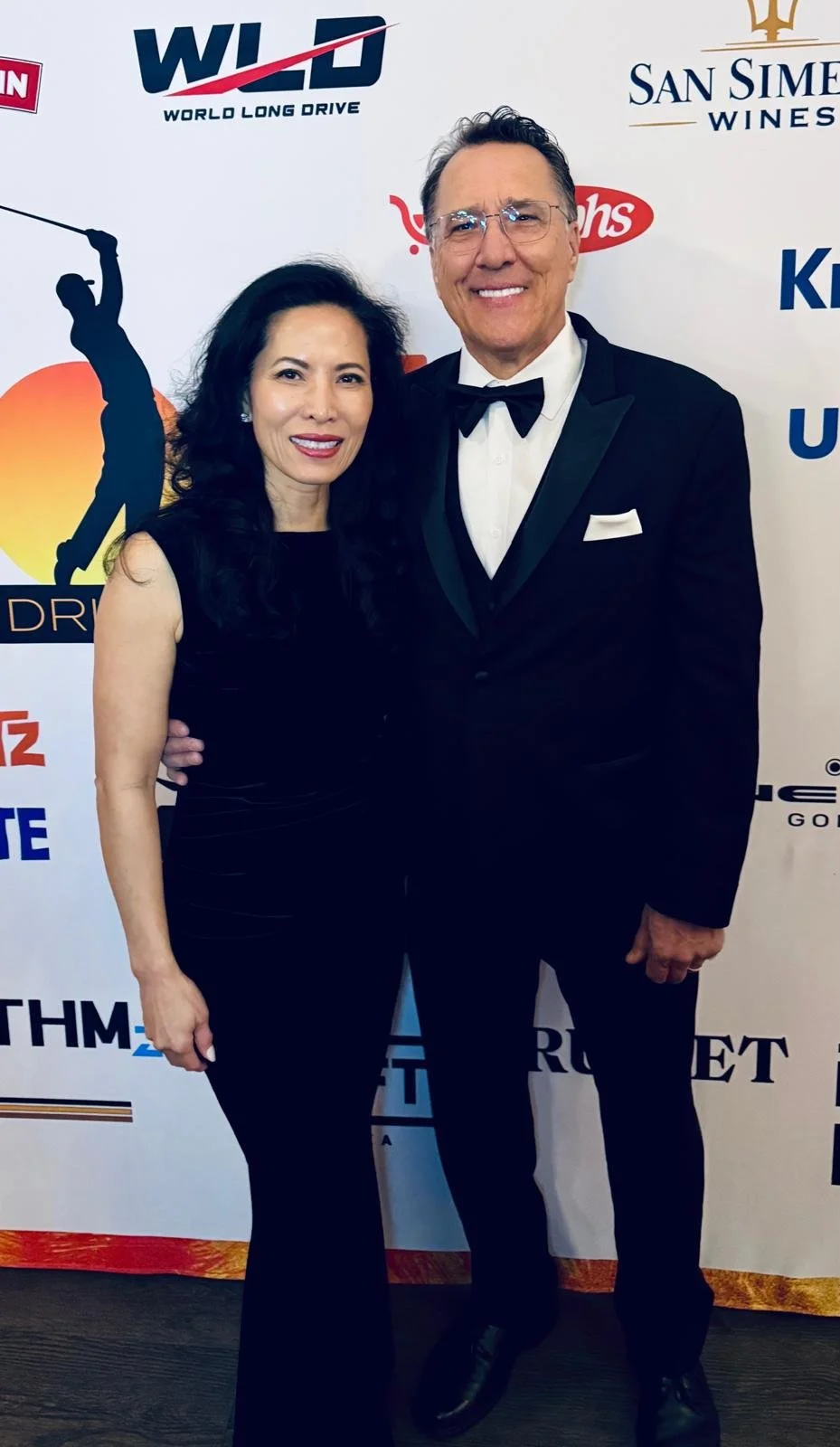 Kimberly and husband dressed in formal attire posing together at an event with a step and repeat banner behind them, featuring logos including WILD, San Simeon Wines, and others.