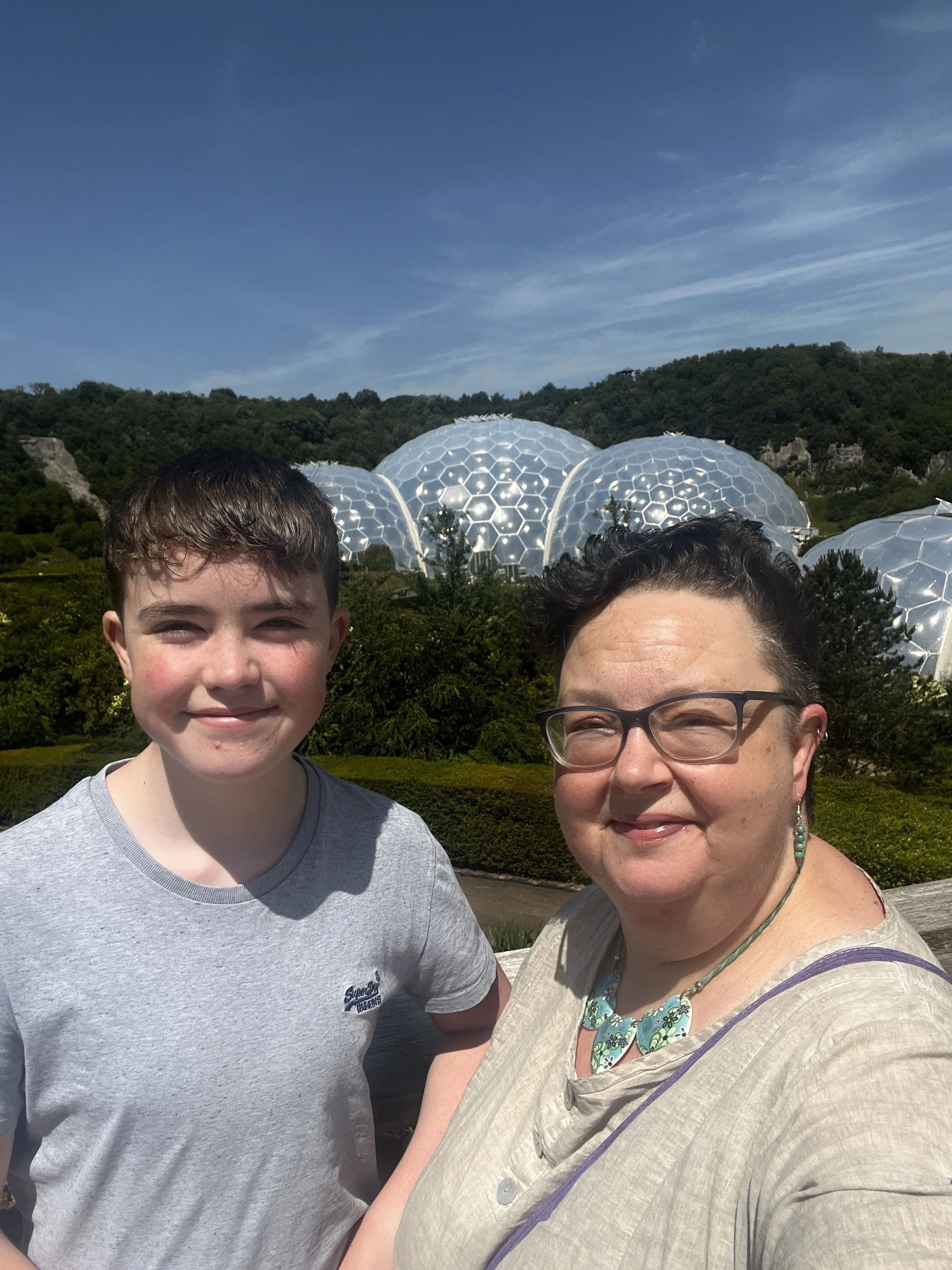 A woman and a young man taking a selfie outdoors with futuristic geodesic dome structures and lush green hills in the background.