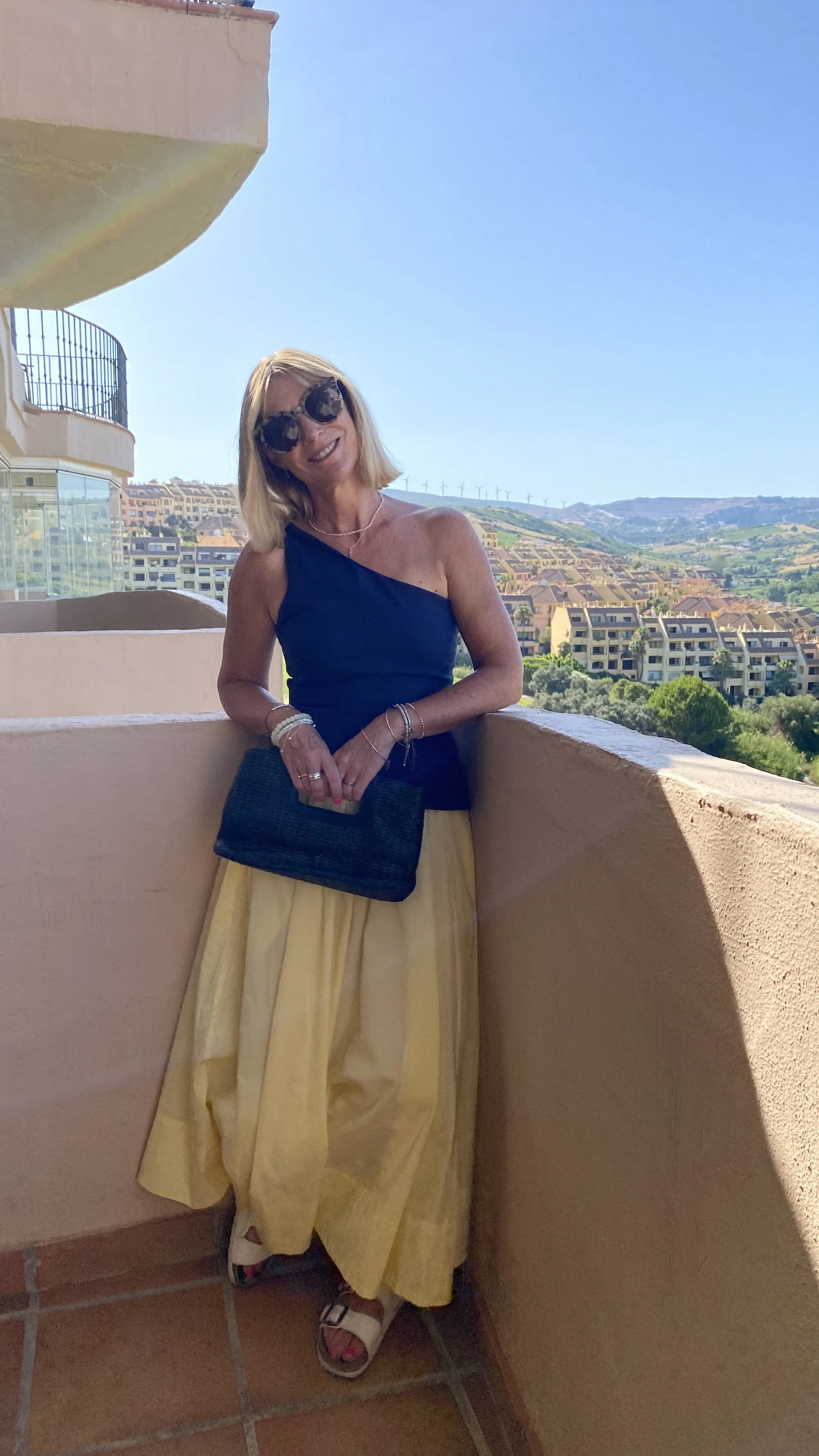 Photo of a glamorous Natasha, with a one sleeves top and bright yellow skirt, on a balcony with a sunny backdrop.
