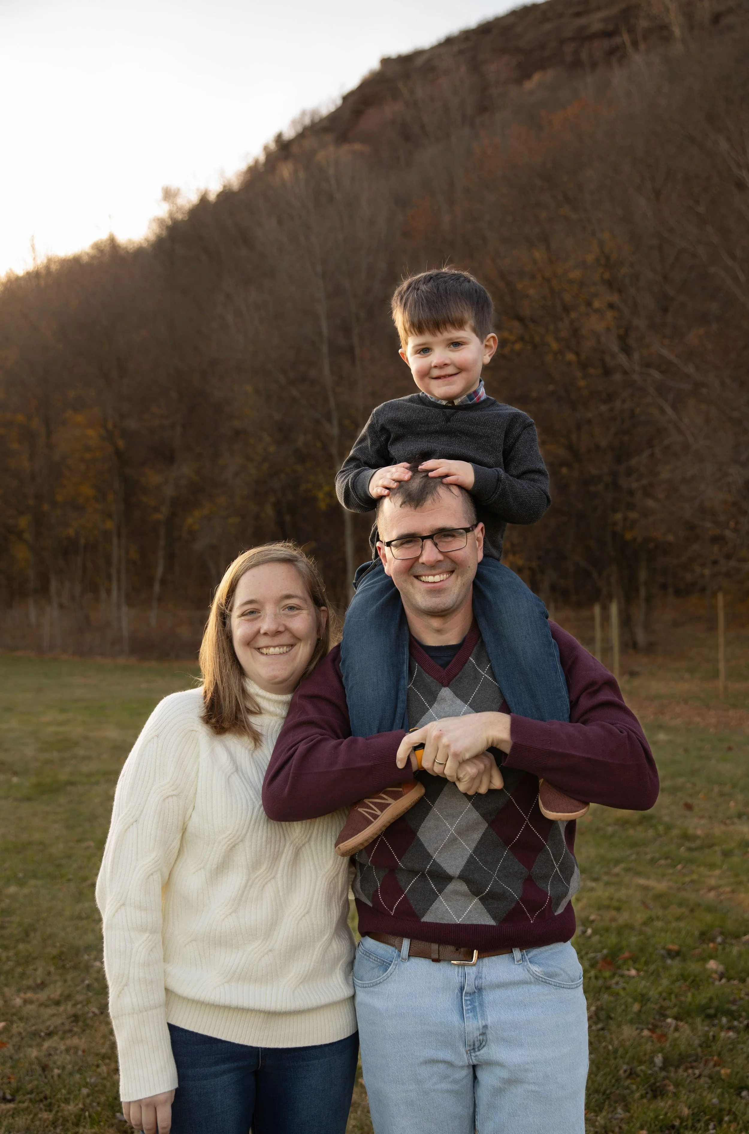 Loryn next to her husband who is carrying their young son on his shoulders. Both smiling wearing jumpers for cold weather.