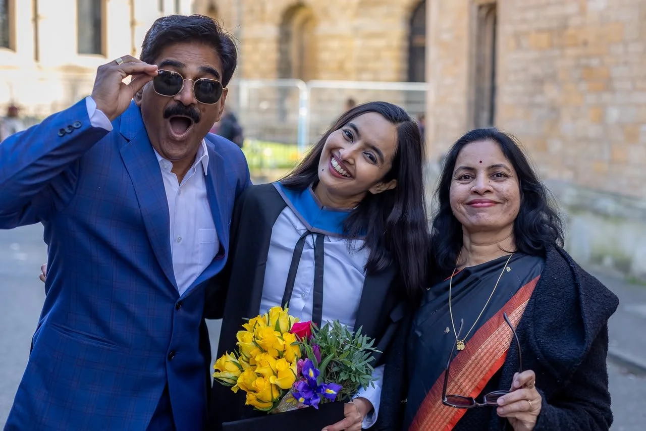 Raskia wearing a graduation gown, holding bright yellow flowers, with her parents wearing formal clothes, all smiling. dad pulling down sun glasses and funny open mouth expression