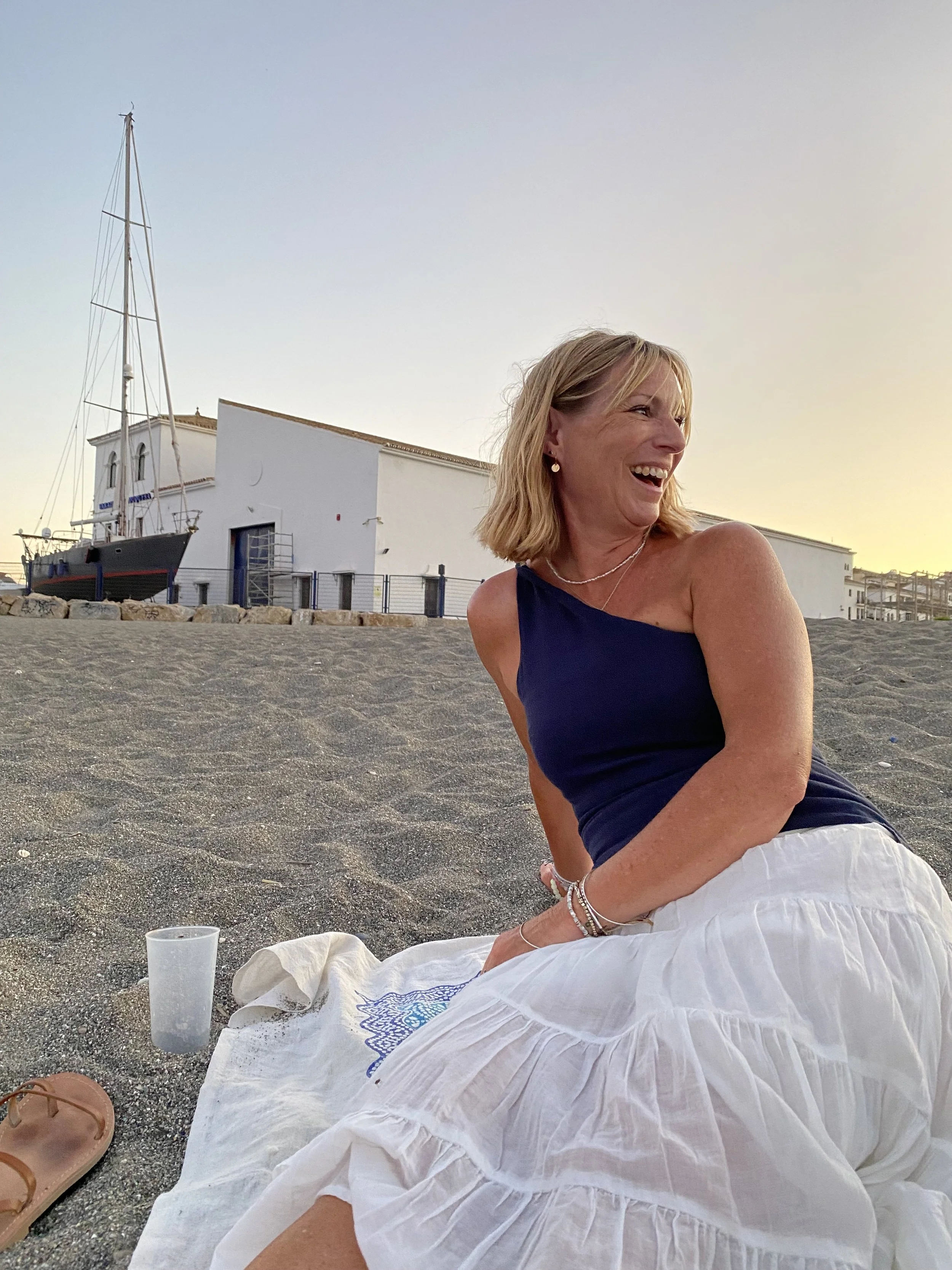 Photo of a smiling Natasha in a one shoulder top, white skirt, laying on a sandy beach with a sail boat and boat house in the backdrop.