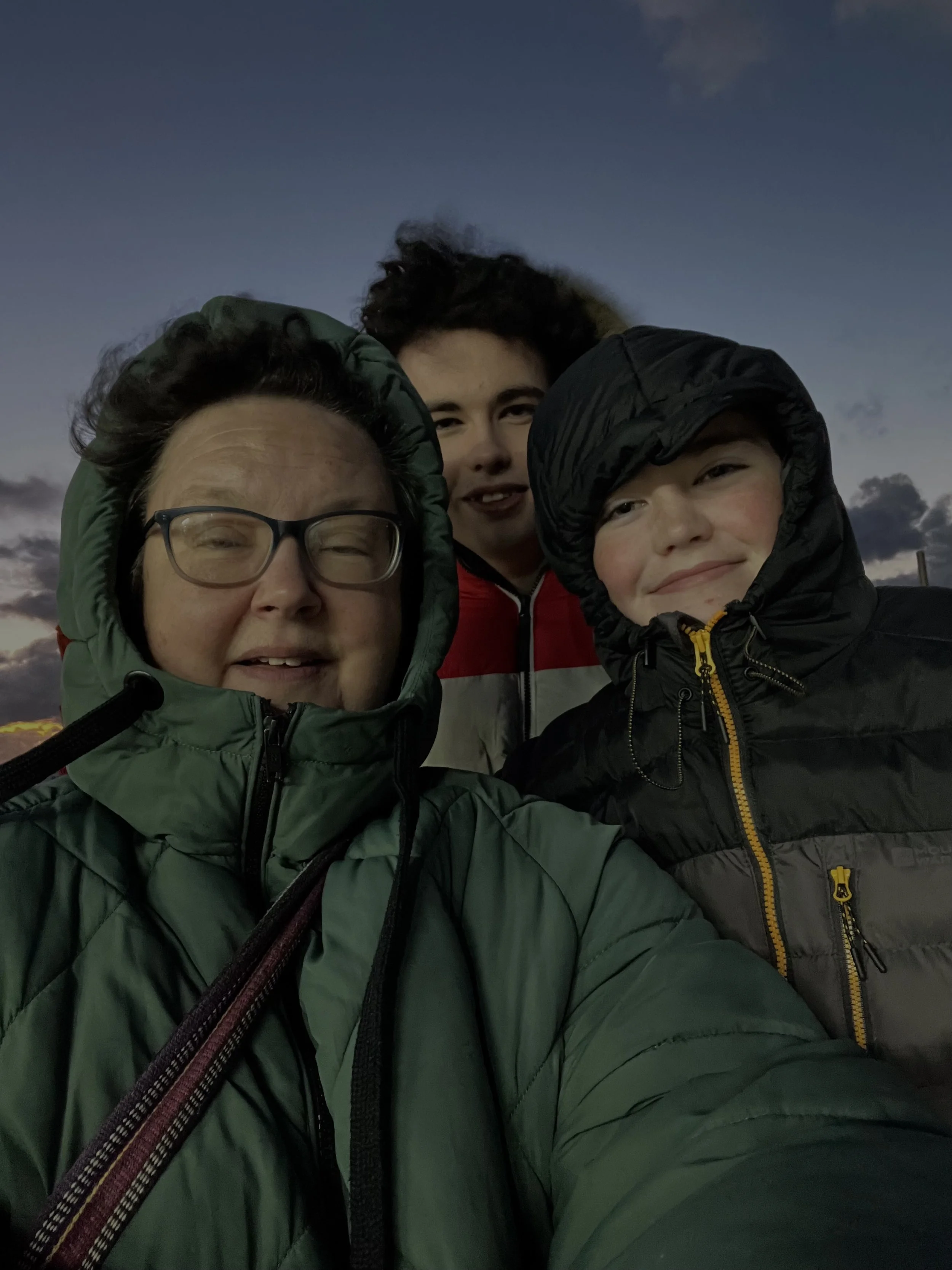 Close-up of three people outdoors during dusk, wearing warm jackets and hoods, smiling and taking a selfie