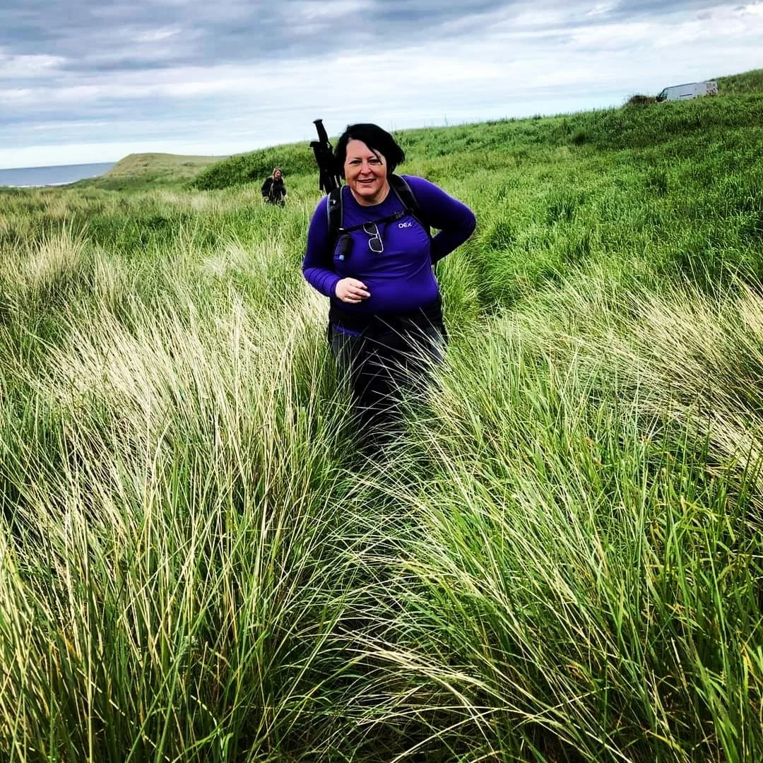 Julia - living with stage 4 ROS1 lung cancer, hiking in a field of tall grass. She has hiking polls and athletic wear and is smiling