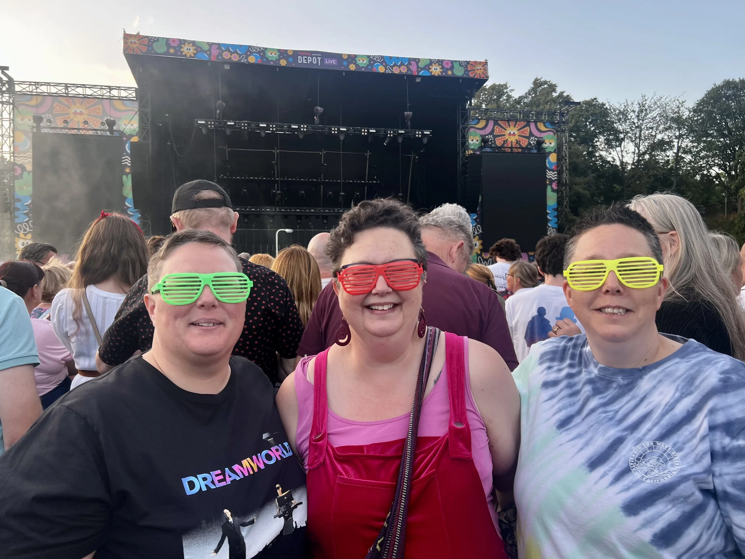 A group of people at an outdoor event or concert, wearing colorful shutter sunglasses, with a stage in the background decorated with vibrant, colorful patterns.