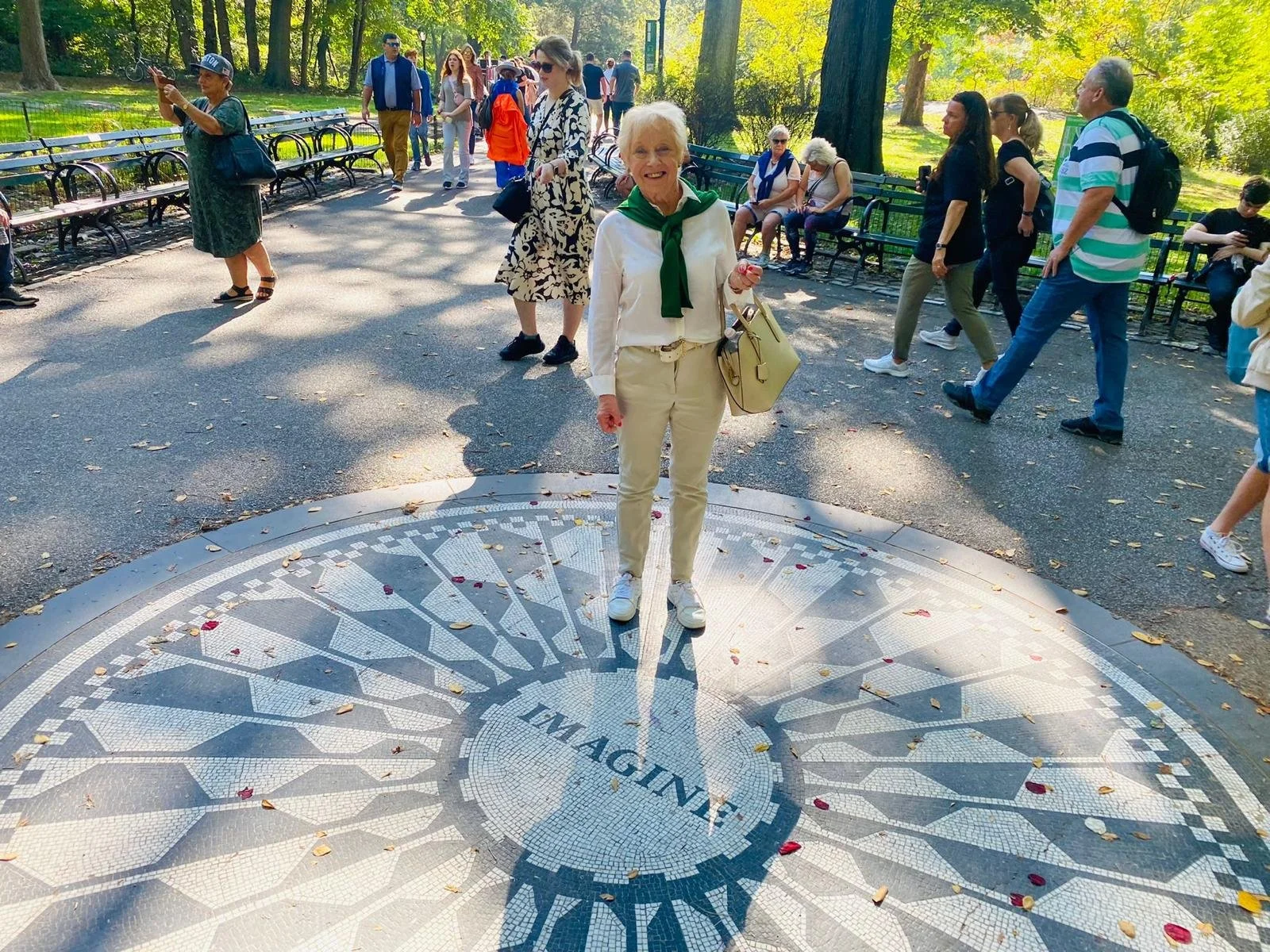 Jenny smiling, wearing bright yellow jeans, with a green jumper over her shoulders standing on a tile image on the ground outside in a park that says  "imagine". She was told there was no hope.