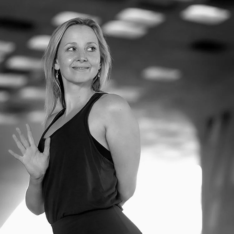 Eine lächelnde Frau in einem schwarzen Top, die sich dreht und die Hand hebt, im Hintergrund eine moderne, helle Raumdecke.