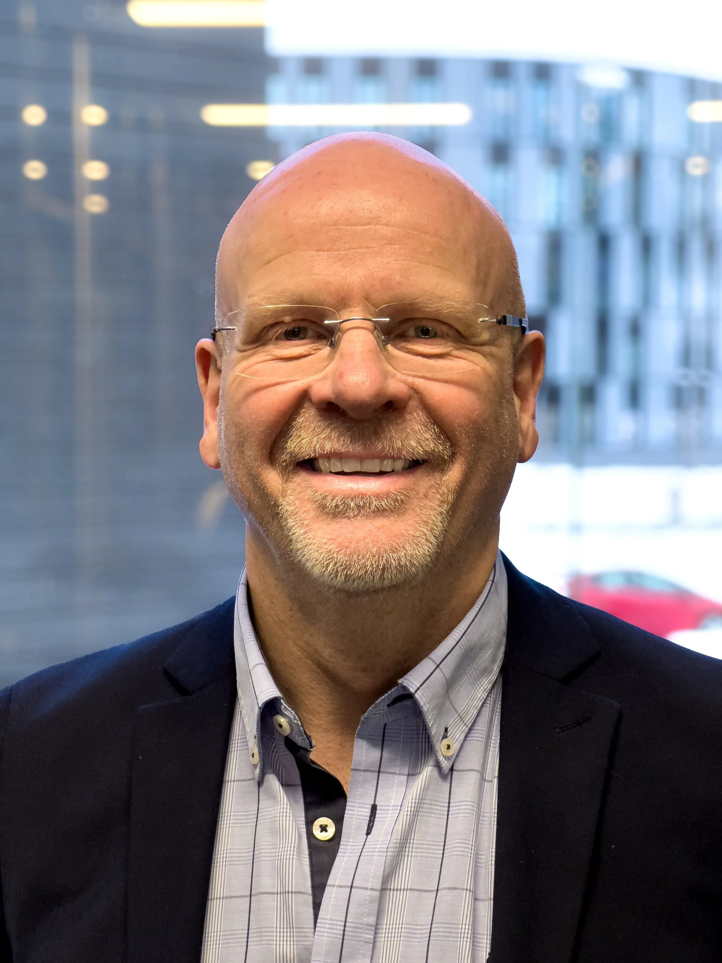 Portrett av en smilende, eldre mann med brillene på nesetippen, i dress og skjorte, foran et kontorvindu.