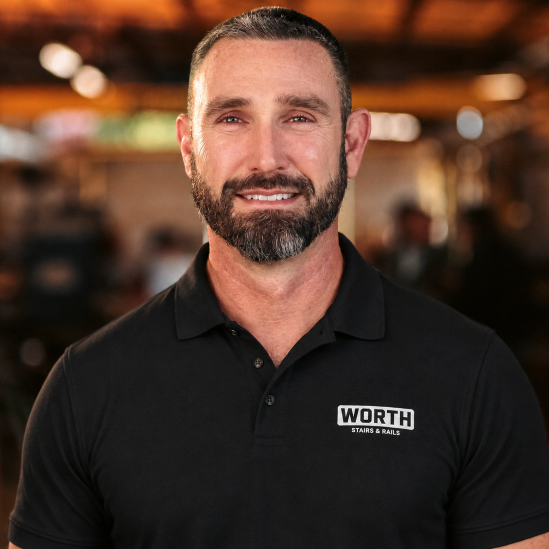 John Combs, Estimator of Worth Stairs & Rails. A middle-aged man with a beard and short hair wearing a black shirt, smiling in an indoor setting with warm lighting and blurred background.