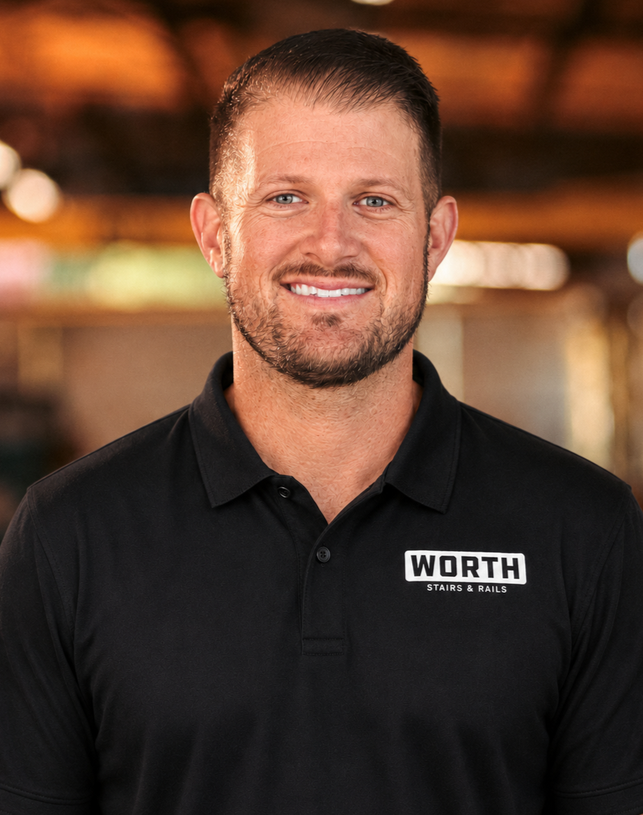 Caleb Worth, VP of Worth Stairs & Rails. A smiling man with short brown hair and facial hair wearing a light blue checkered button-up shirt, standing indoors with a blurred background of warm lighting and wooden elements.