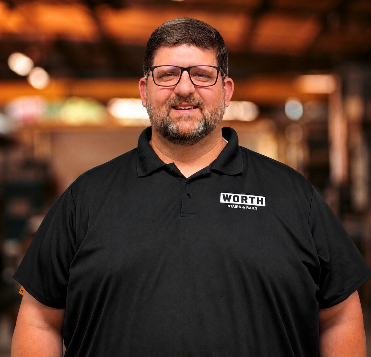Tommy Carter, Project Manager of Worth Stairs & Rails. A man with a beard and glasses smiling, wearing a light gray polo shirt, in an indoor setting with warm lighting and blurred background.