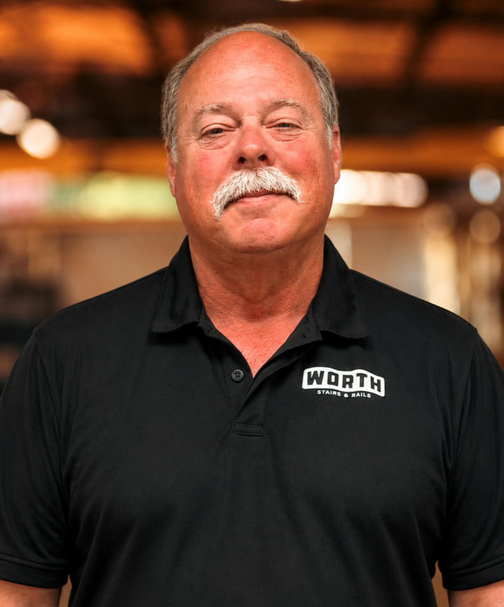 Tim Worth, President of Worth Stairs & Rails. A man with a moustache and gray hair smiling, wearing a blue Hawaiian shirt with a white octopus pattern, standing indoors with a blurred background of warm lighting.