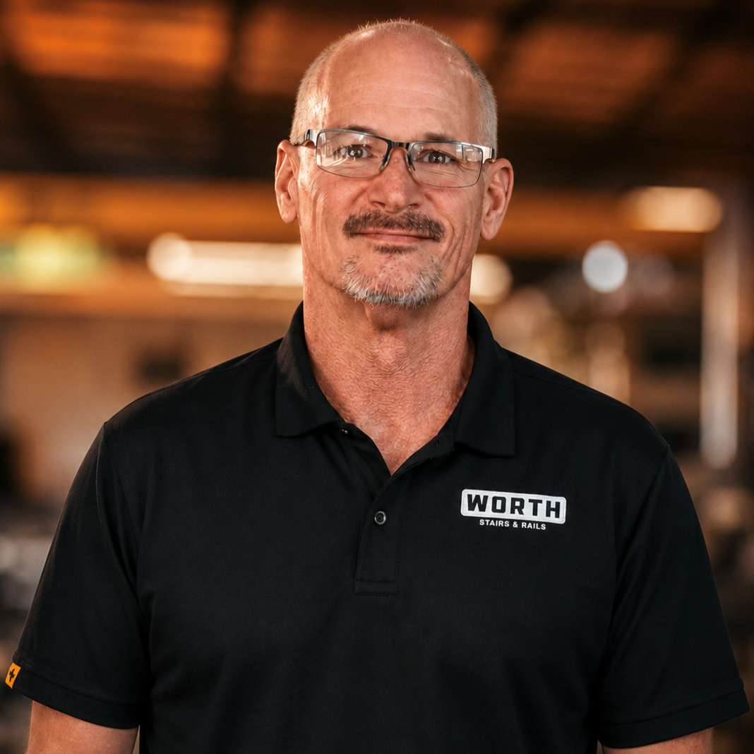 Todd Mathews, Detailer of Worth Stairs & Rails. A middle-aged man with short gray hair, glasses, and a goatee, smiling while wearing a black collared shirt with a flag patch on the sleeve, inside a warmly lit indoor space.