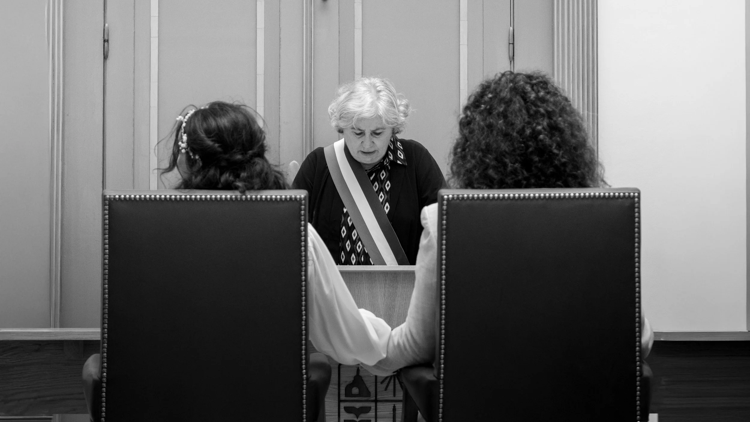 Deux femmes sont assises face à face à une table, et une femme âgée se tient derrière la table, regardant vers le bas, dans un environnement intérieur.