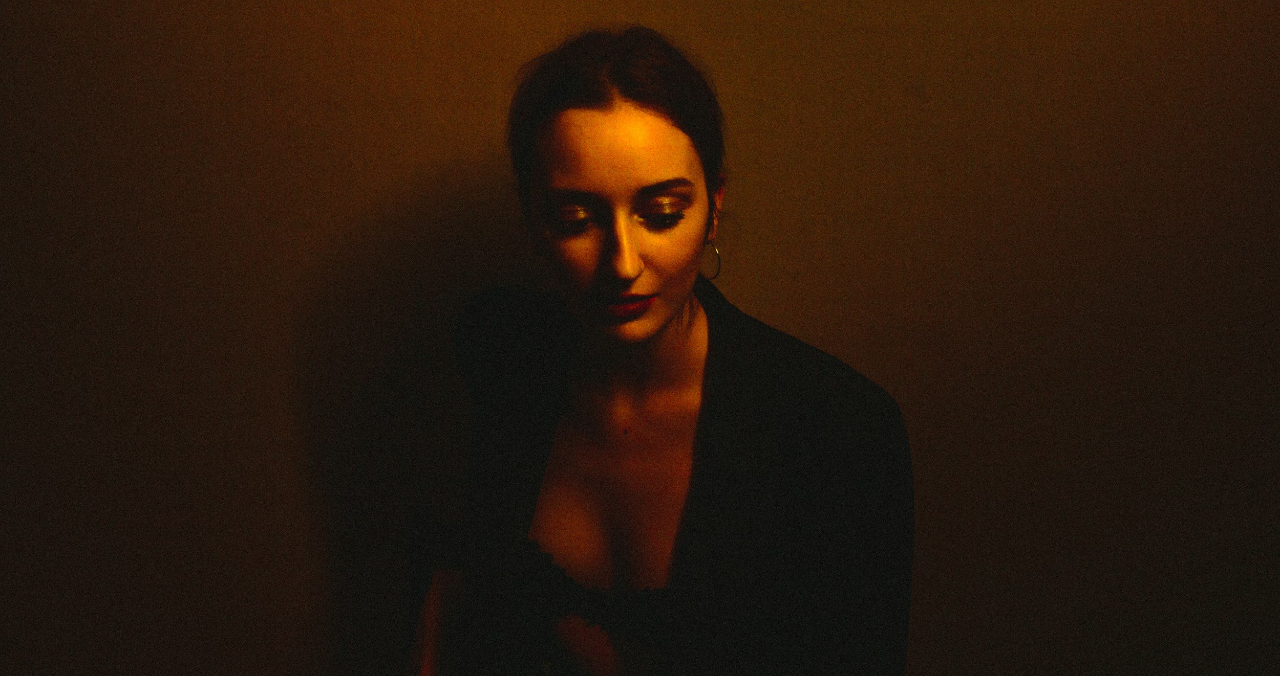 Low-light portrait of a woman with dark hair pulled back, wearing a dark jacket and hoop earrings, looking downward under warm, moody lighting against a dark, textured background