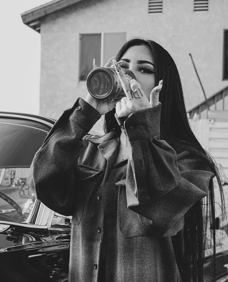 Black-and-white photo of a light-skinned woman with long dark hair wearing an oversized jacket and drinking something while flipping the camera off with the other hand