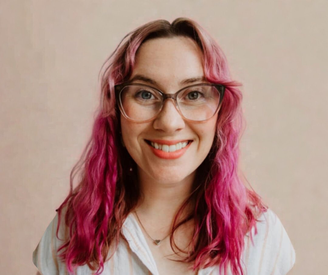 A woman with pink, curly hair and glasses smiling at the camera against a plain beige background.