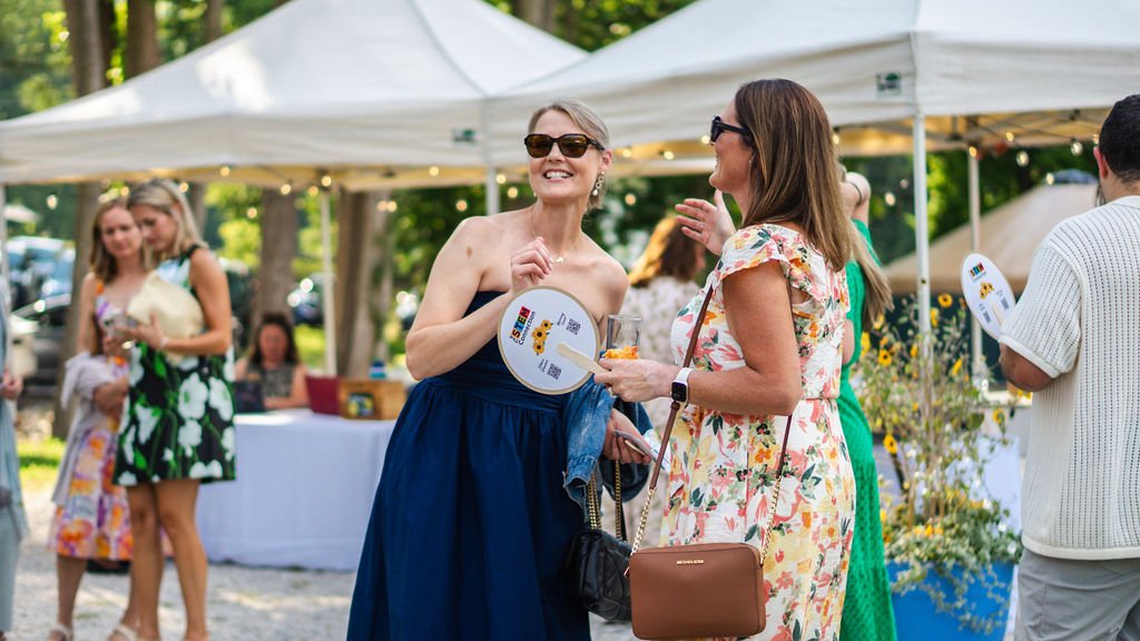 Two women in summer dresses and sunglasses smiling and talking outdoors at a festive event with white tents and string lights.