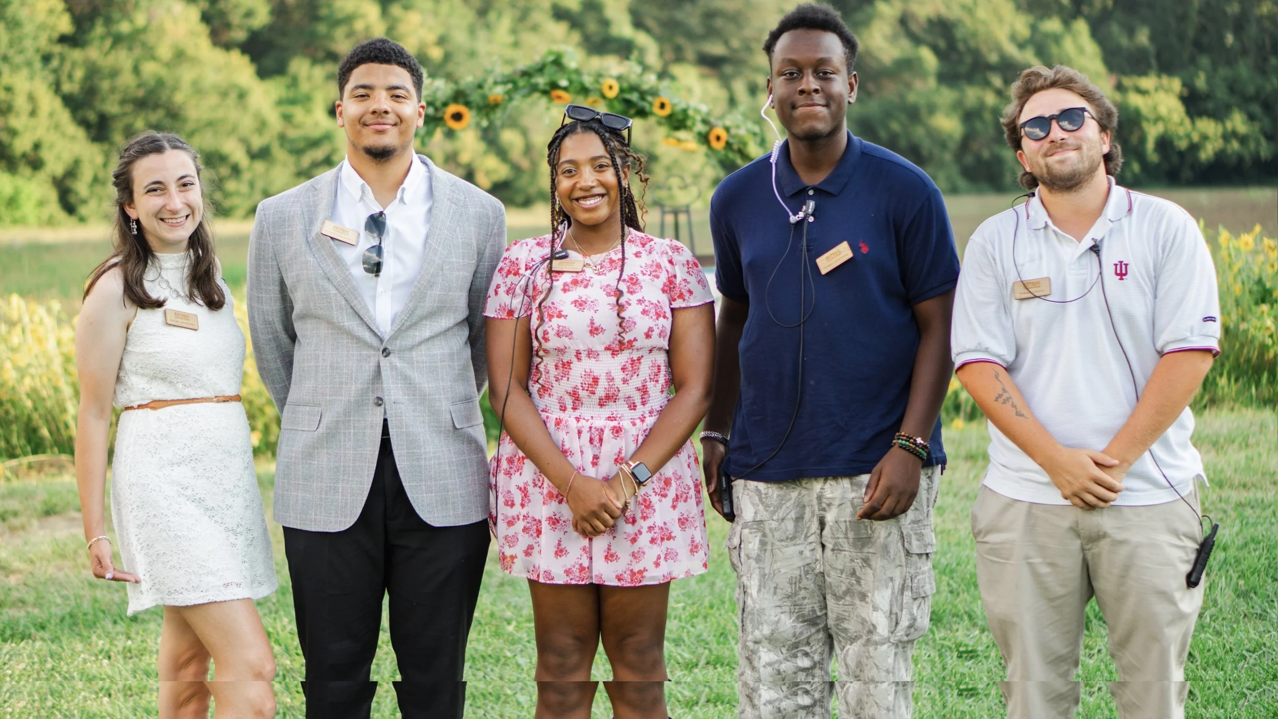 Group of five young adults standing outdoors in a park, smiling at the camera, with trees and greenery in the background.