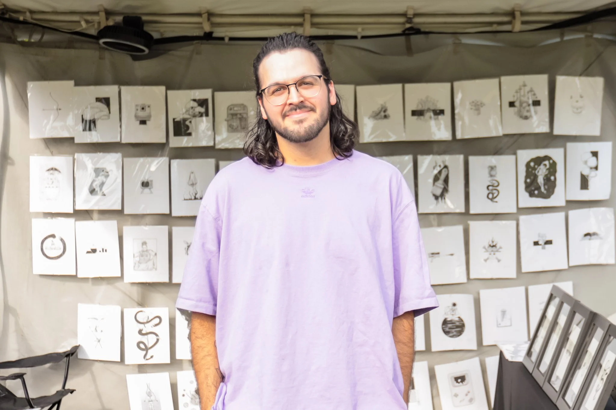 Young man with glasses and long hair wearing a purple T-shirt, standing in front of an art display of black and white drawings on paper attached to a wall.