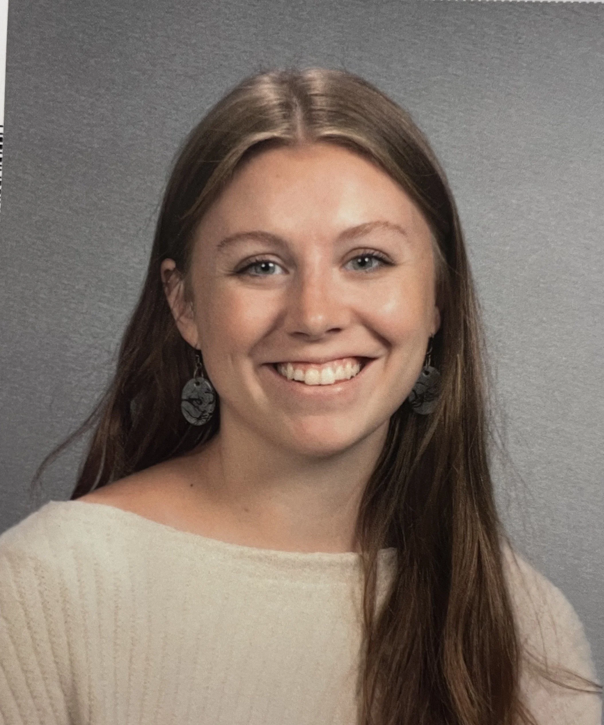 A woman with long brown hair smiling, wearing black earrings and a cream-colored sweater, against a gray background.