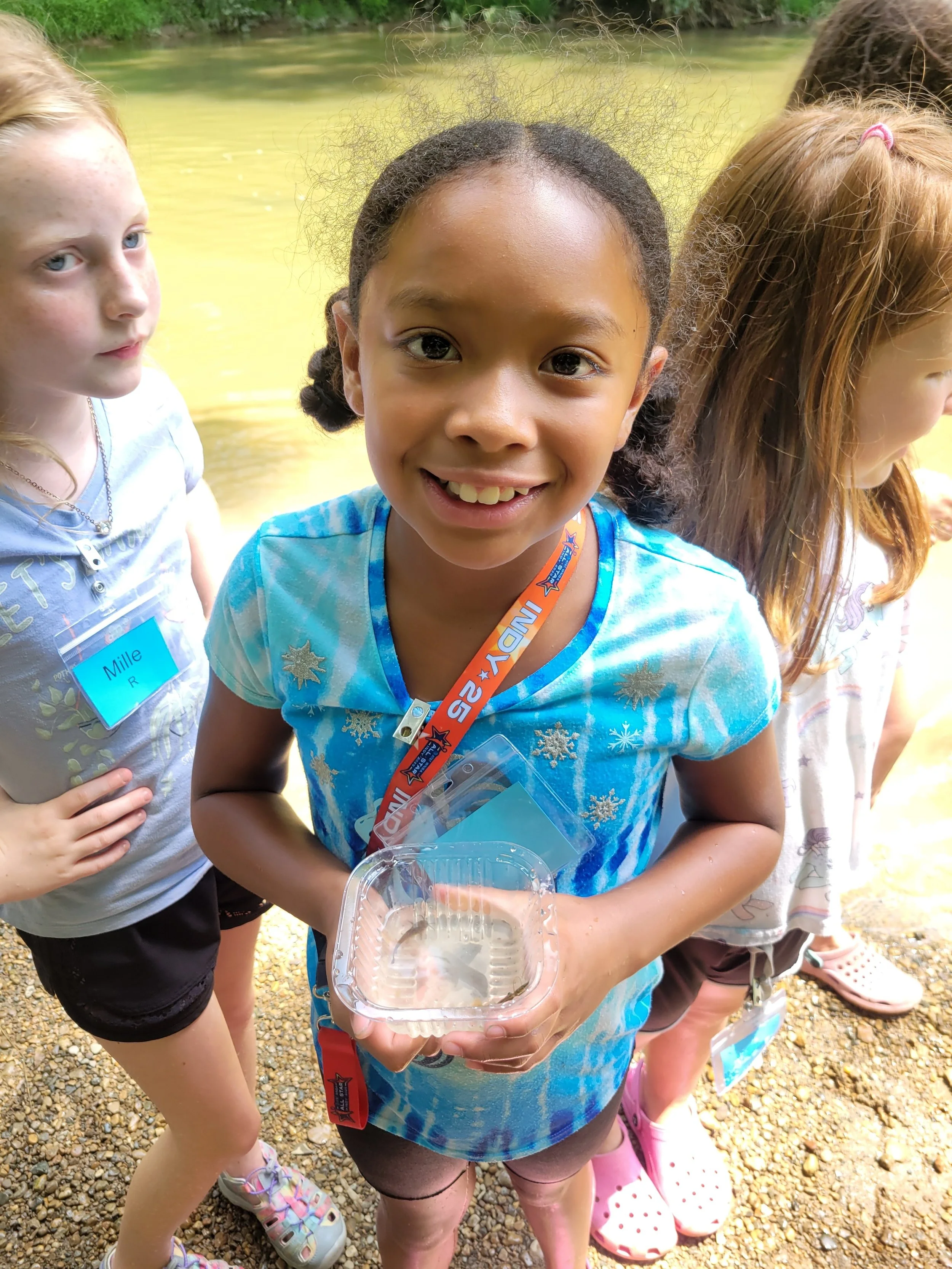 Smiling young girl holding a clear plastic container outdoors near water, with two girls partially visible beside her, standing on a gravel surface.