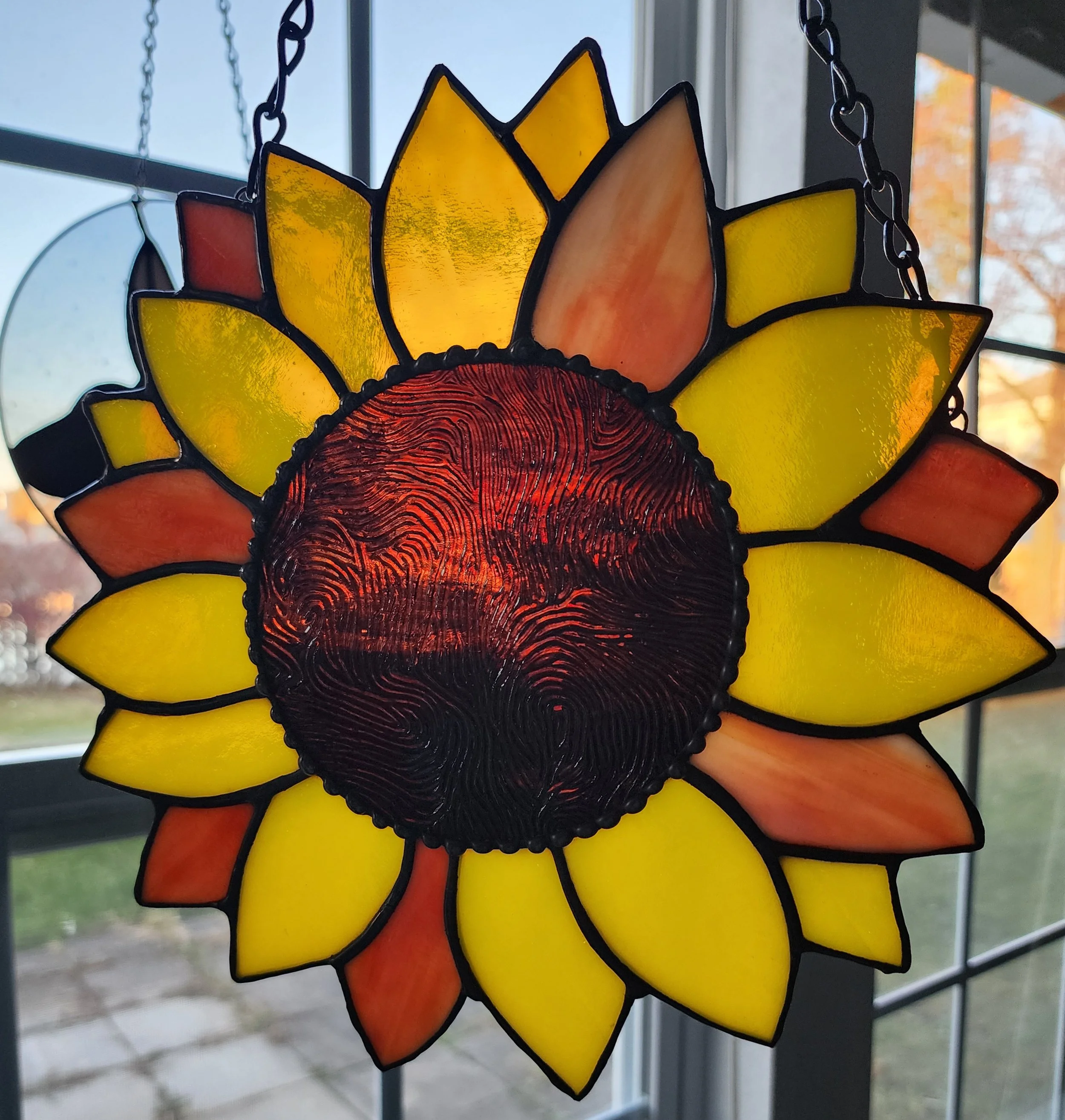 Colorful stained glass sunflower hanging decoration with yellow, orange, and red petal sections, backlit from within.