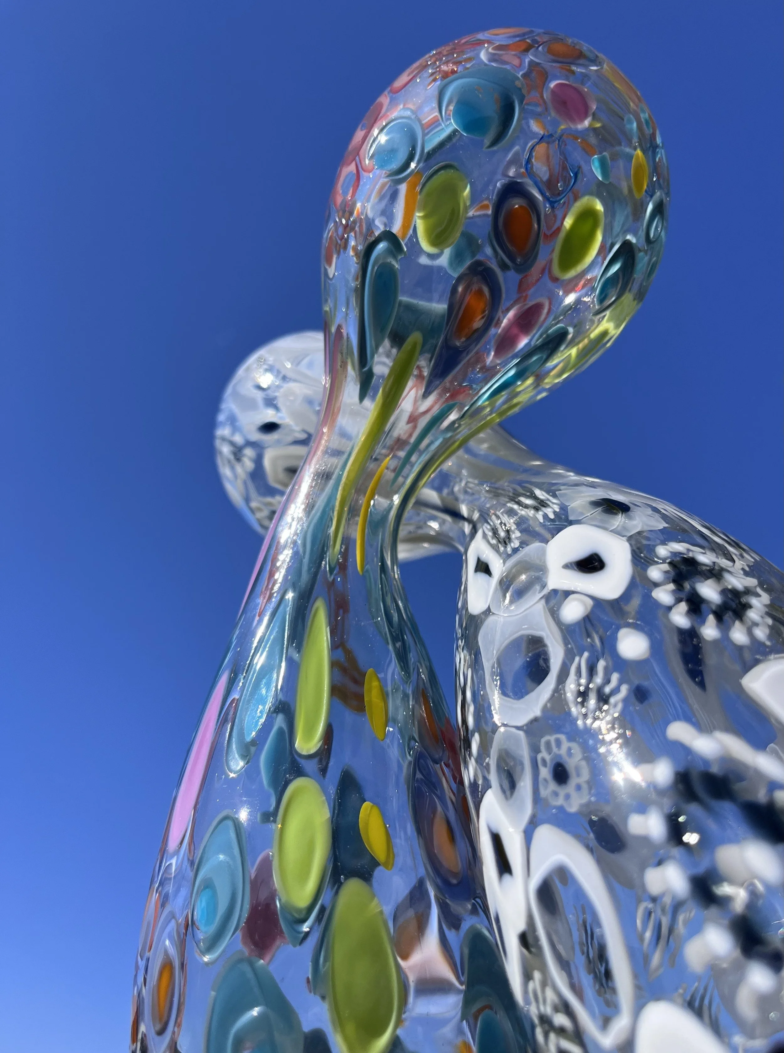 Close-up of a colorful glass sculpture against a clear blue sky, featuring flowing curves and multicolored patterns.