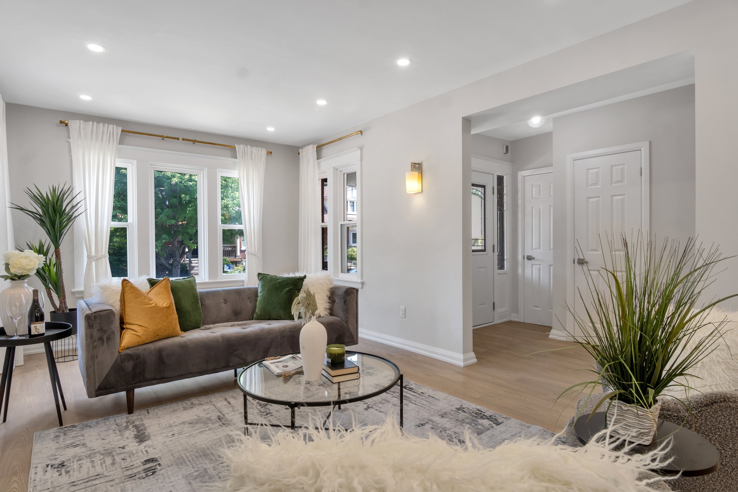 Bright living room with gray sofa, colorful cushions, glass coffee table, indoor plants, and large windows with white curtains.