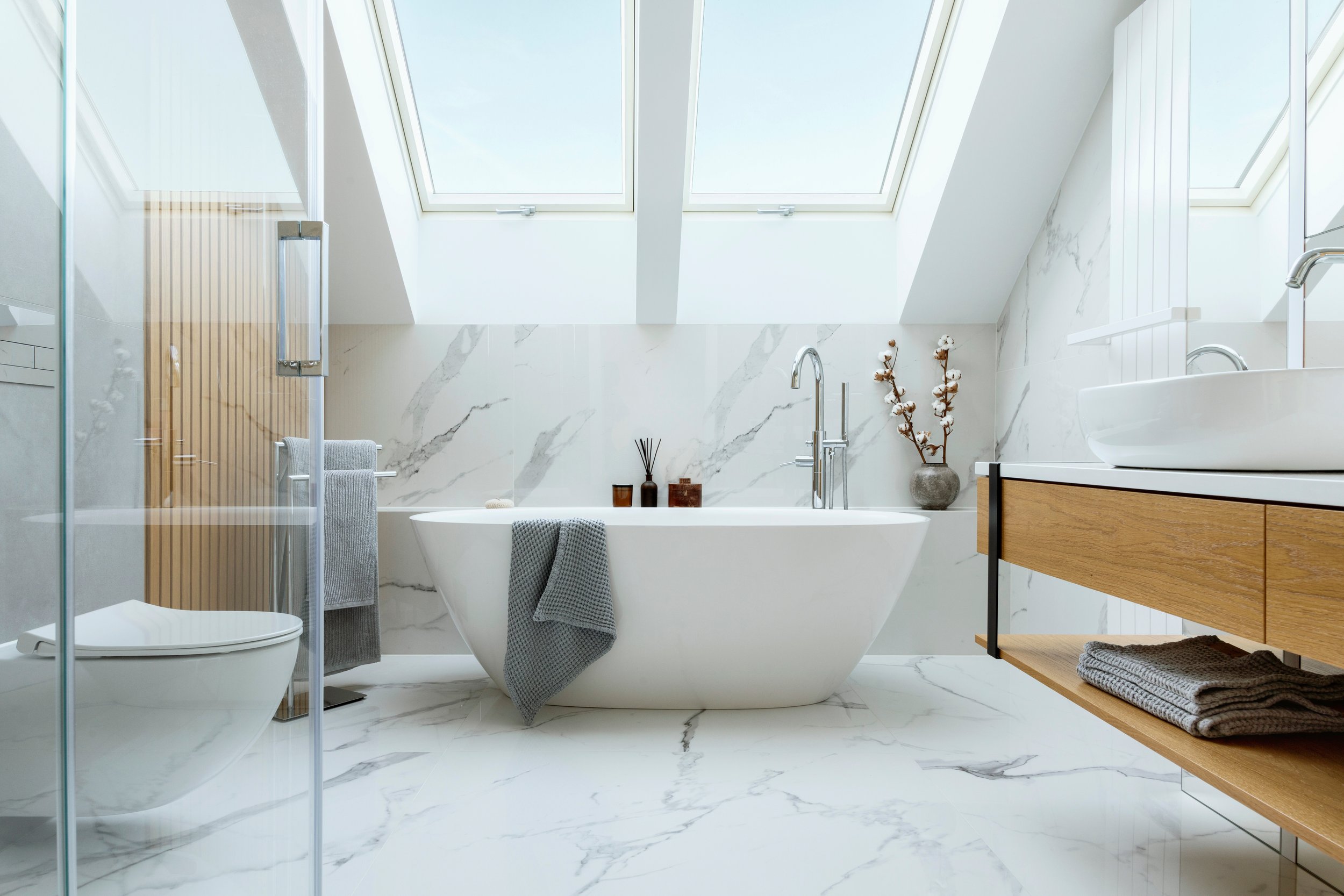 Modern bathroom with freestanding bathtub, skylights, marble walls, wooden vanity, and towels.