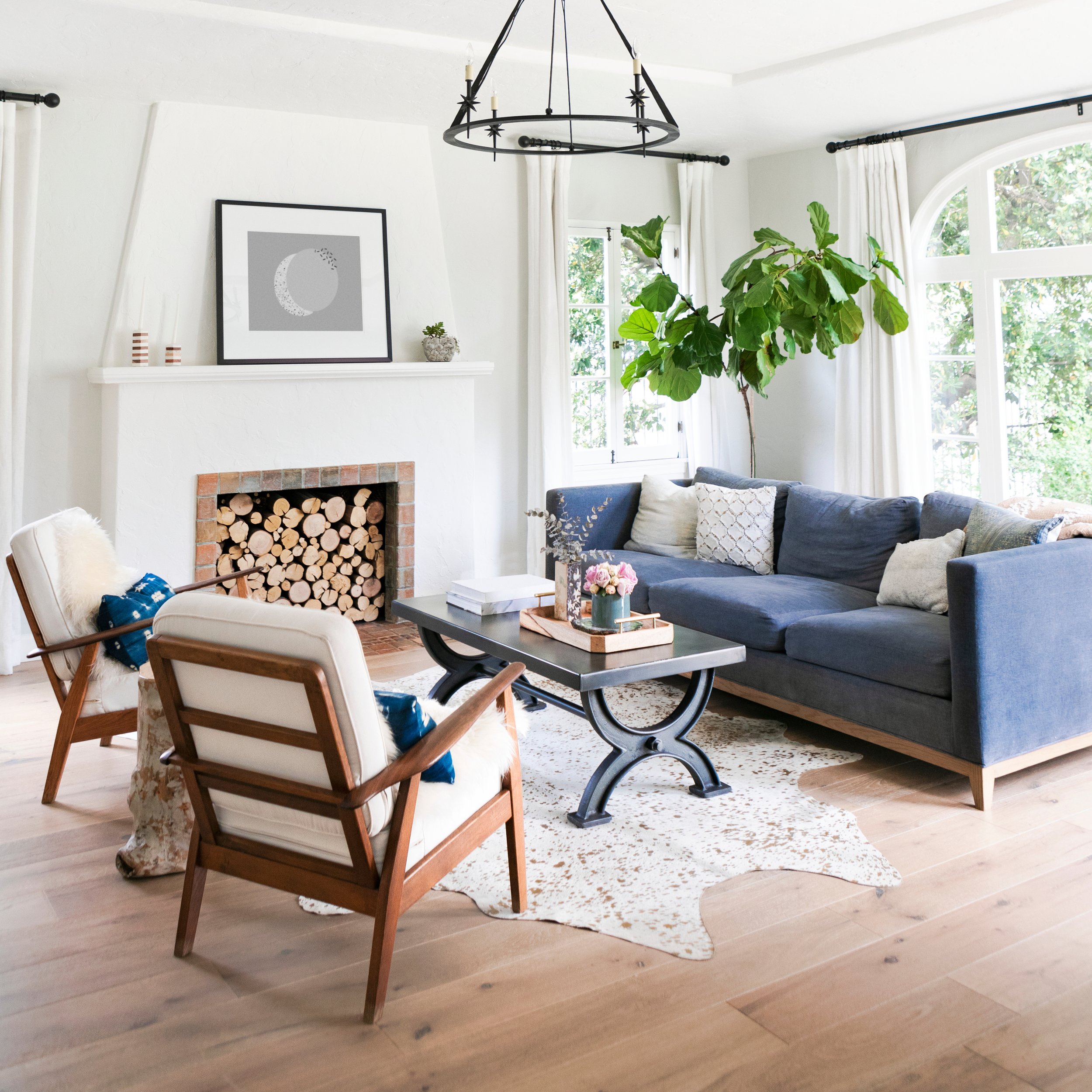 Stylish living room with a navy blue sofa, two wooden armchairs with white cushions, a coffee table, and a white fireplace with stacked logs. A large leafy plant is near the window, and a decorative chandelier hangs from the ceiling.