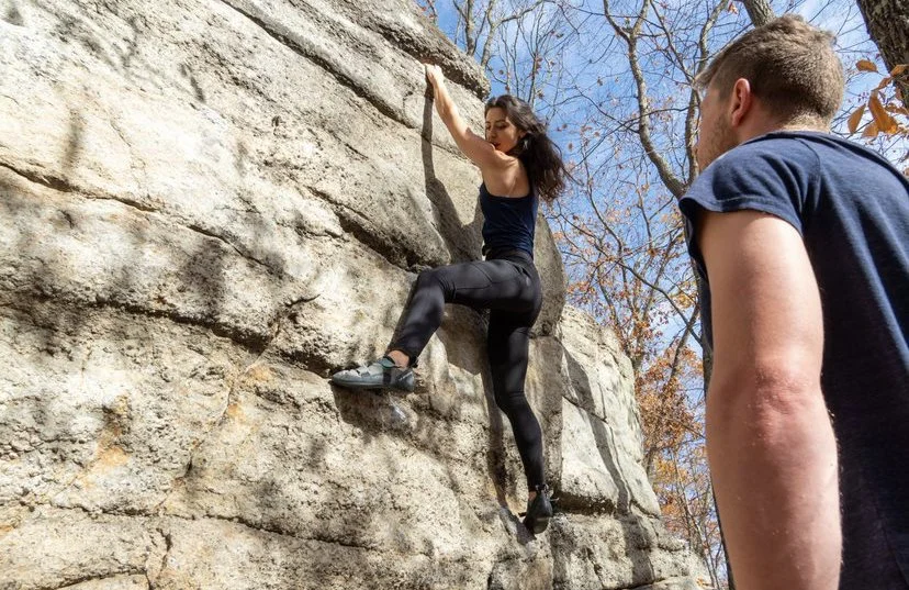 A woman rock climbing outdoors on a nearby rock wall, with a man spotting her from below.